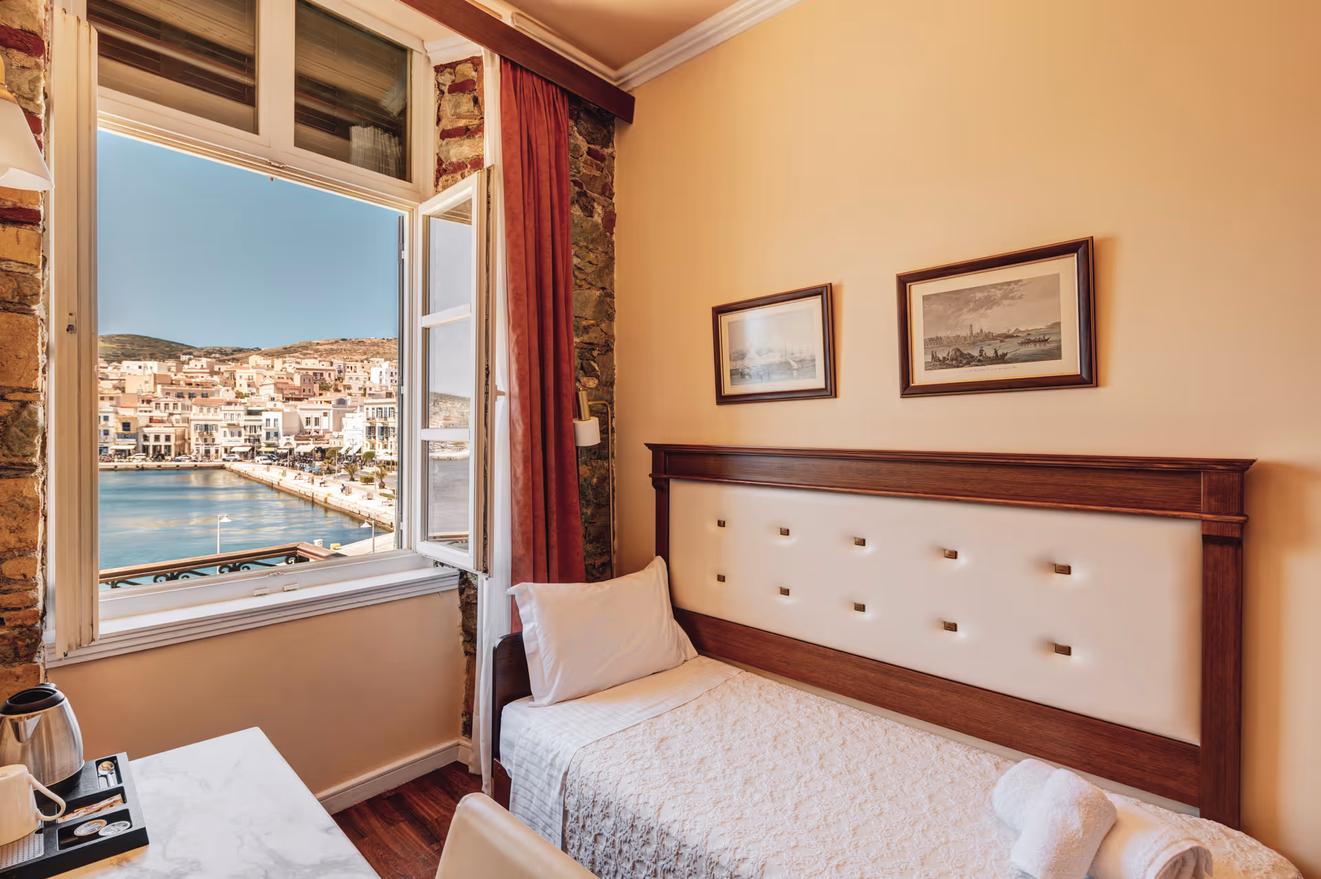 Cozy single bedroom with open window overlooking a waterfront town, featuring a neatly made bed and a small table with a kettle and cups.
