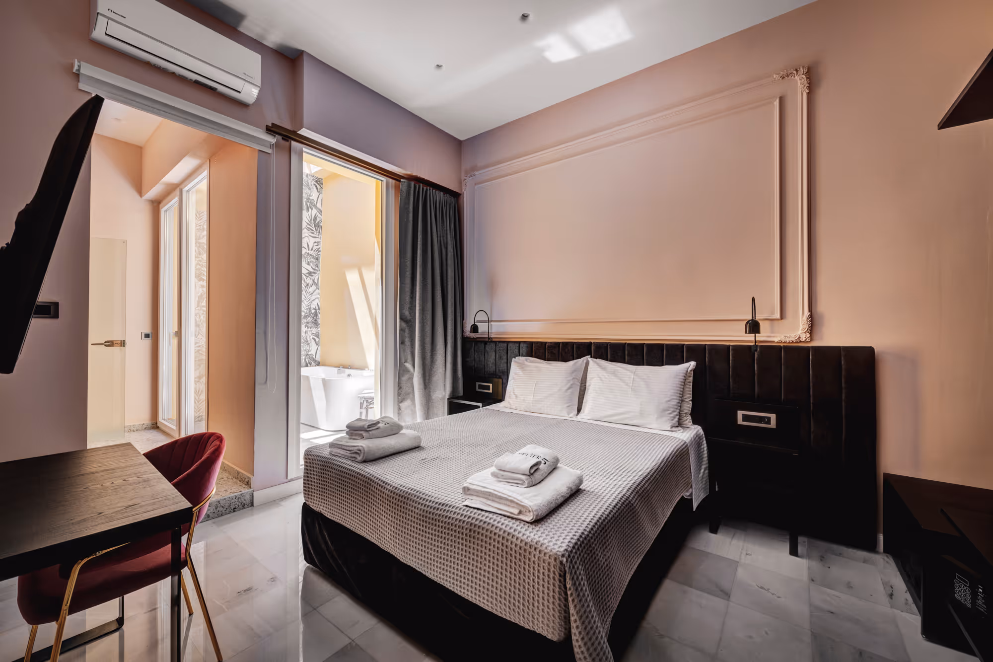 Modern hotel bedroom with a large bed, gray headboard, folded towels, a desk with a red chair, and a bathroom visible through a glass door.