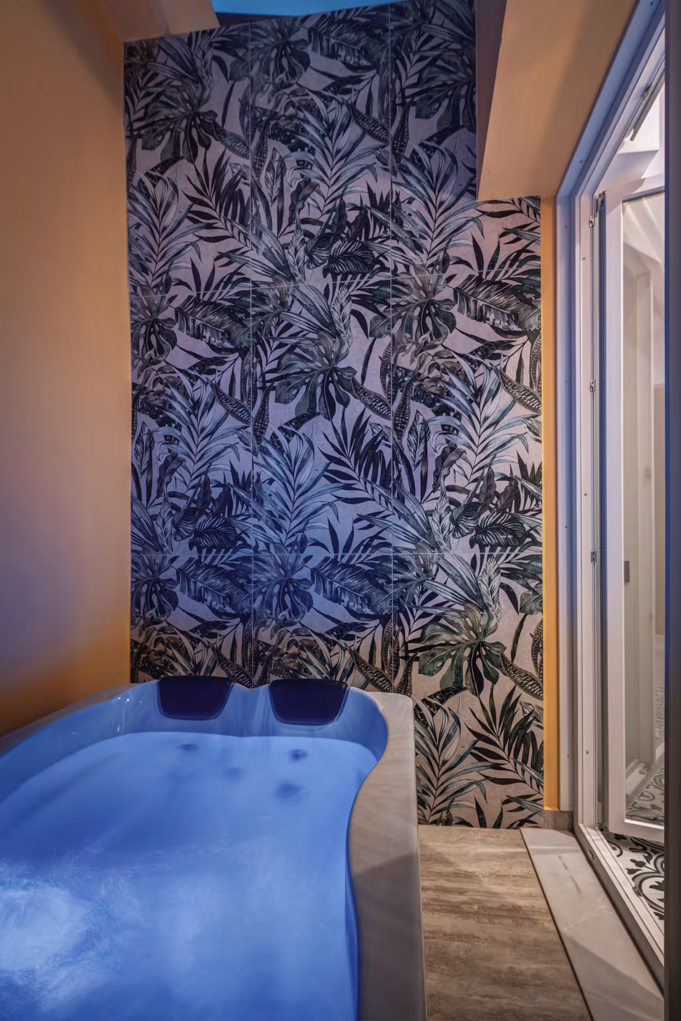 Indoor hot tub with blue water and two headrests, next to a wall with dark leafy tropical wallpaper and a glass door.