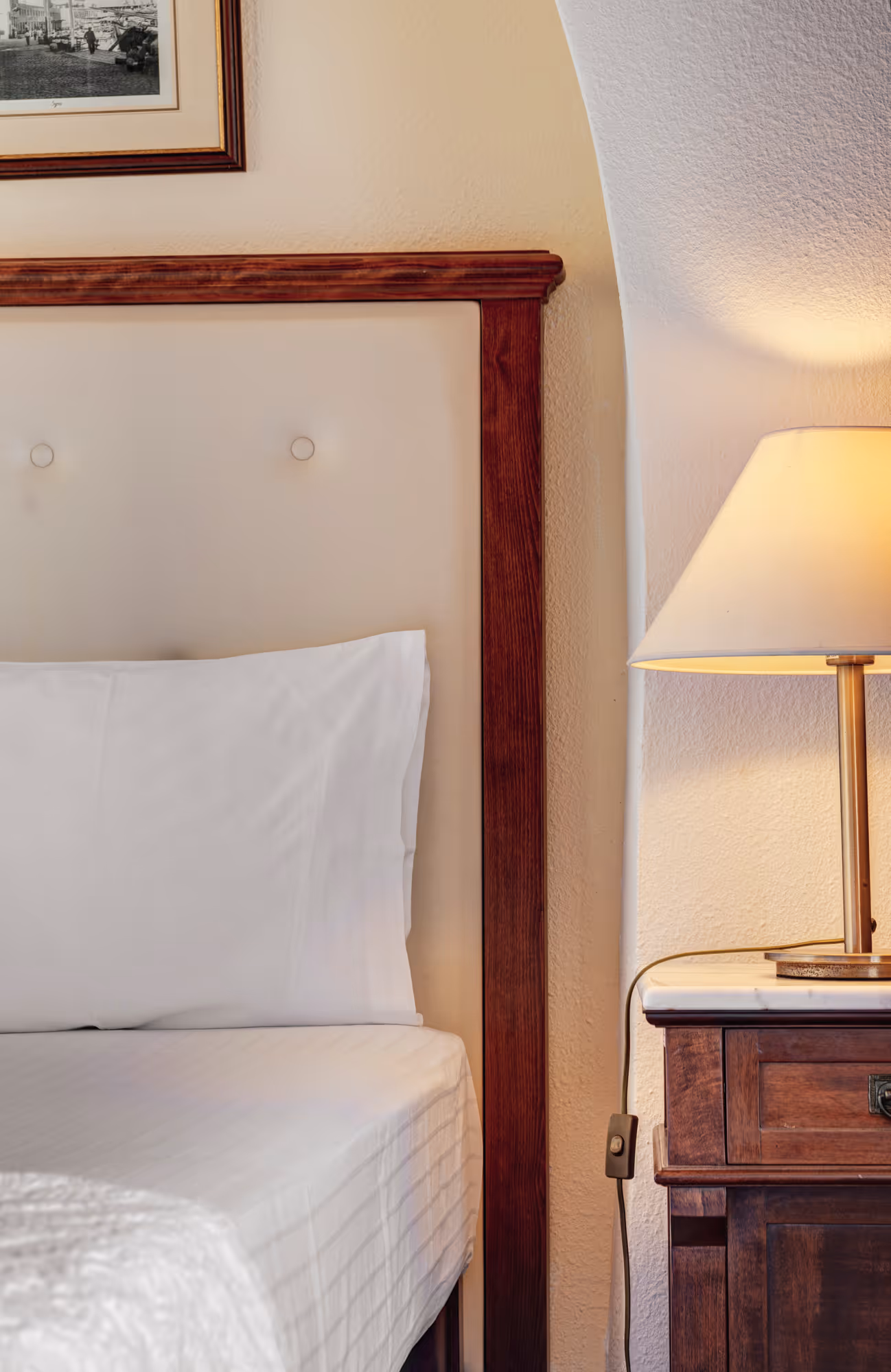 Close-up of a bed with white pillow and bedding, wooden headboard, and a nightstand with a lit lamp.