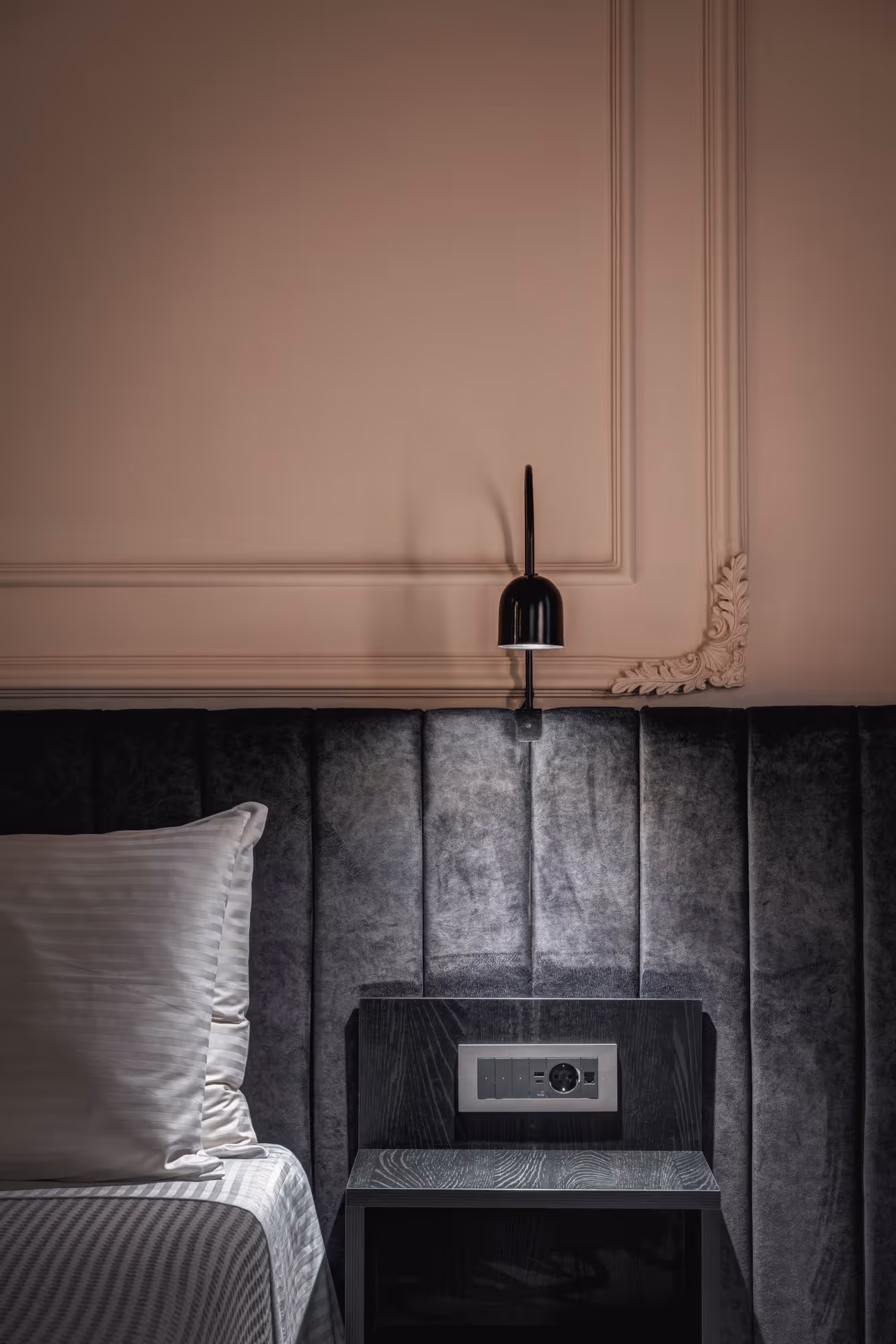 Modern bedroom corner with a grey upholstered headboard, black bedside table with power outlets, and a black wall-mounted lamp.