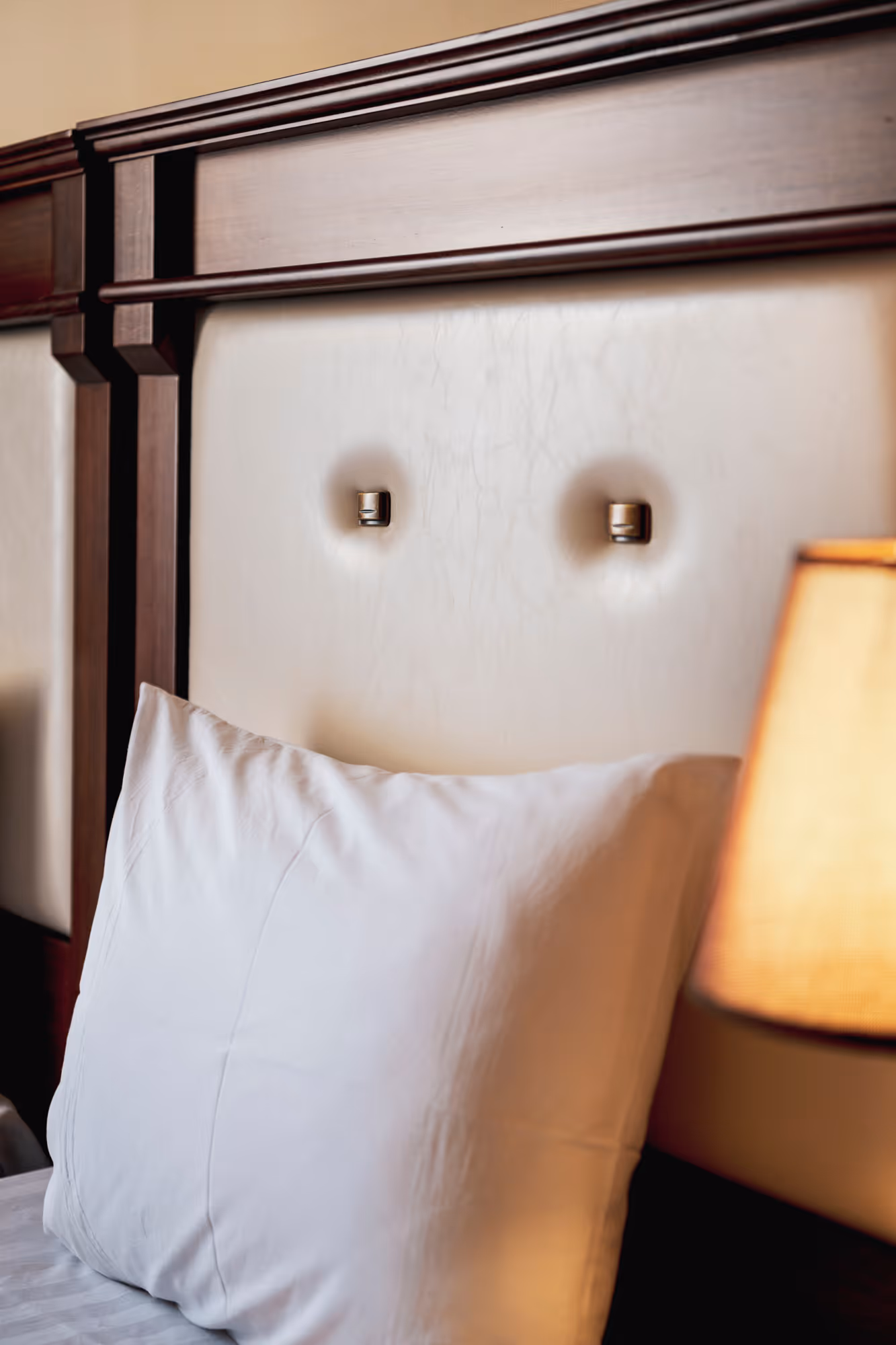 Close-up of a white pillow resting against a cream-colored headboard with decorative wooden trim and a lit bedside lamp.