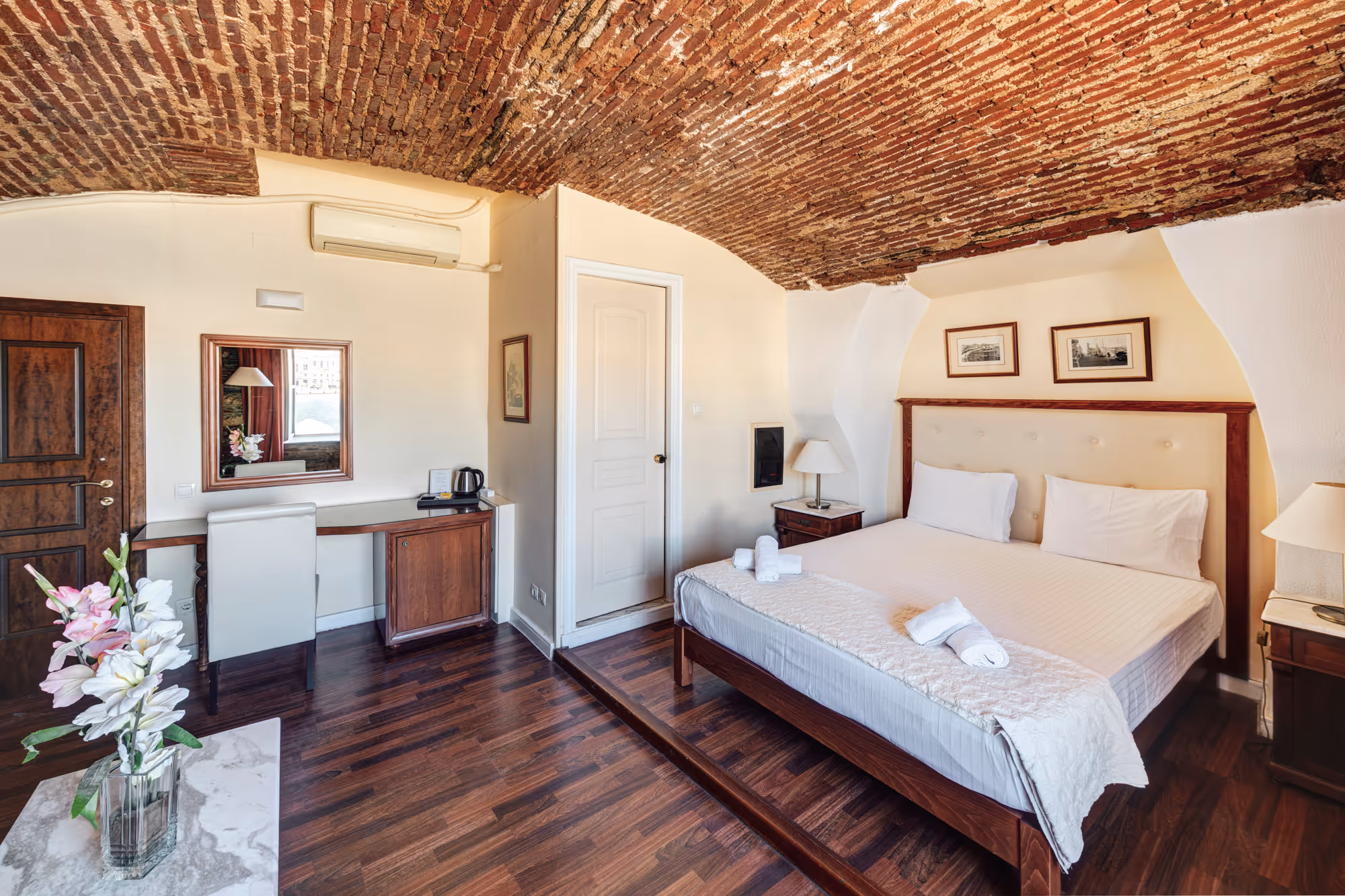 Cozy hotel room with exposed brick arched ceiling, wooden floor, king-size bed with white linens, bedside tables with lamps, desk with chair and mirror, and a vase of flowers on a marble surface.