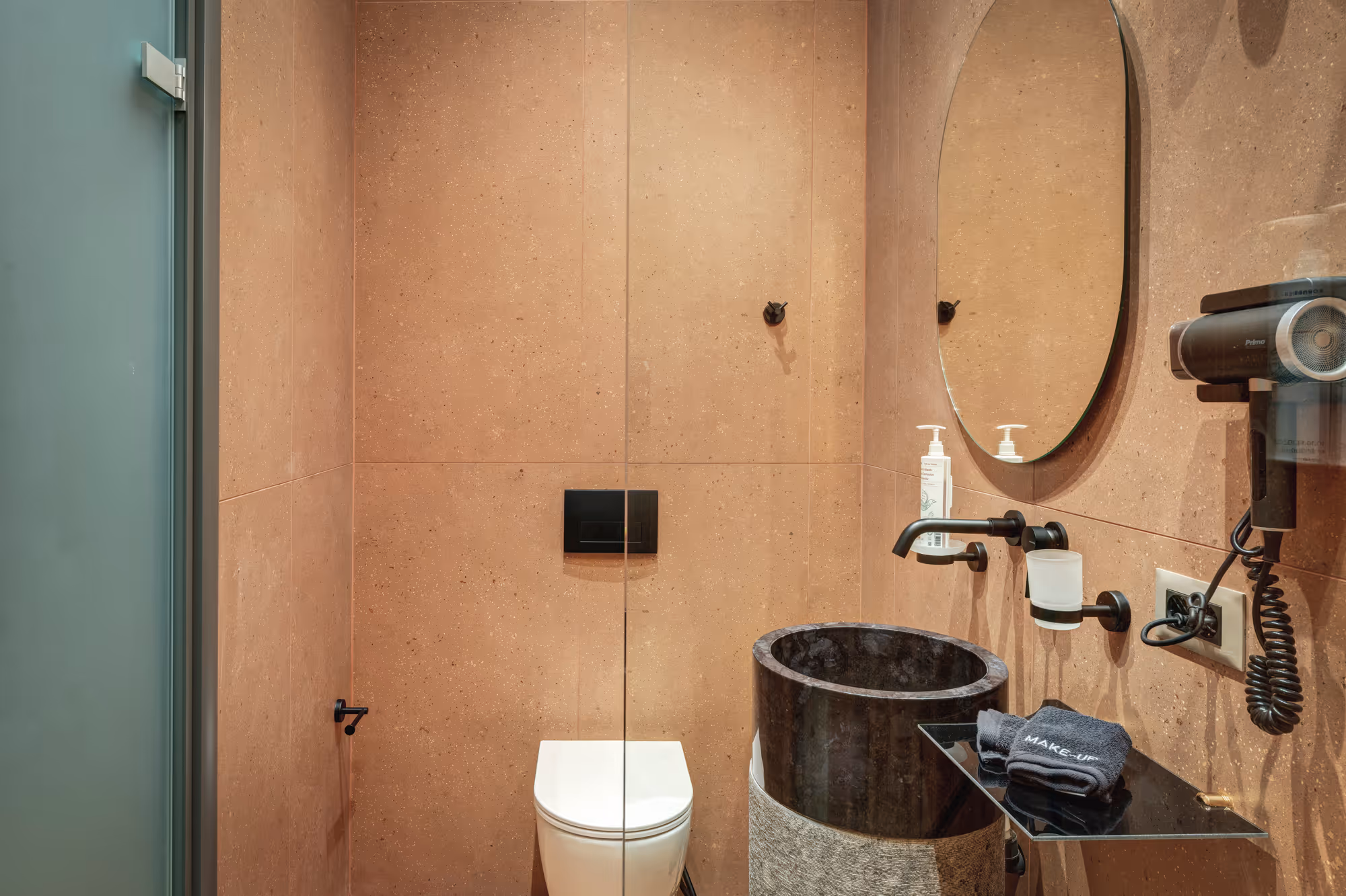 Modern bathroom with beige tiled walls, a white toilet, a round black stone sink, an oval mirror, and a wall-mounted hair dryer.