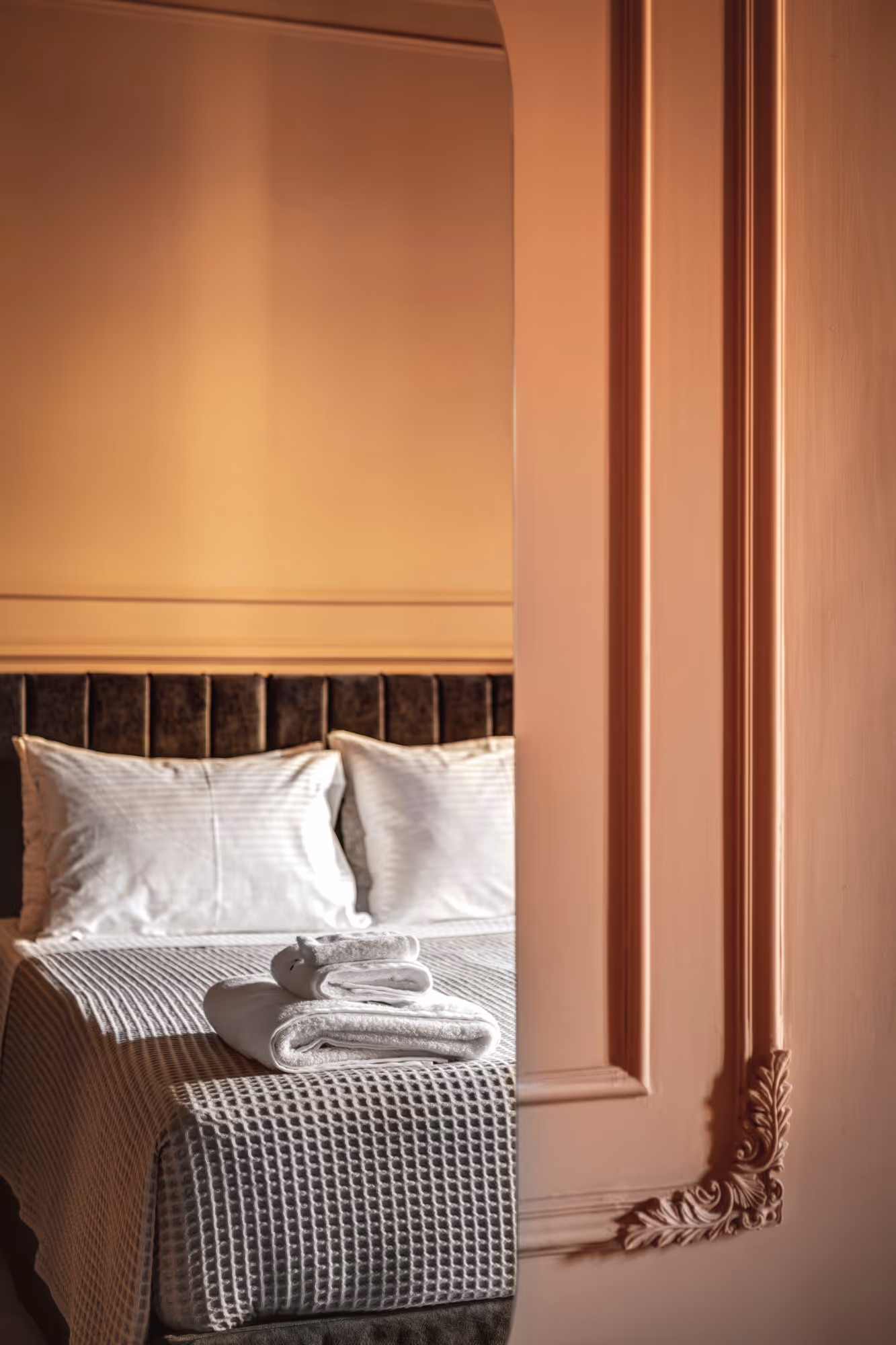 Cozy bed with white pillows and folded towels on a textured grey bedspread, reflected in an ornate peach-colored mirror.