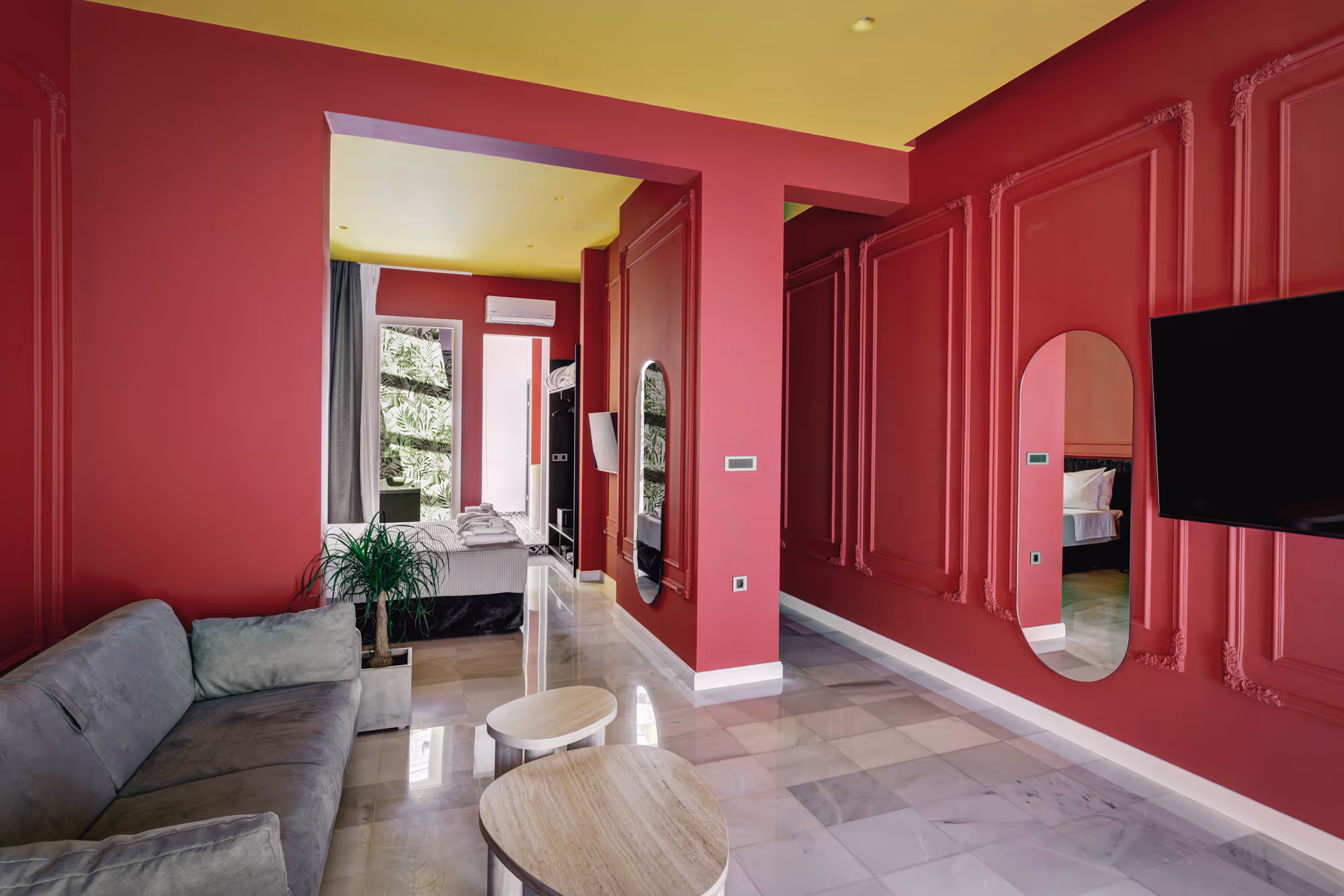 Modern living space with red walls, yellow ceiling, gray sofa, two round wooden tables, and a bedroom with leaf-patterned curtains.