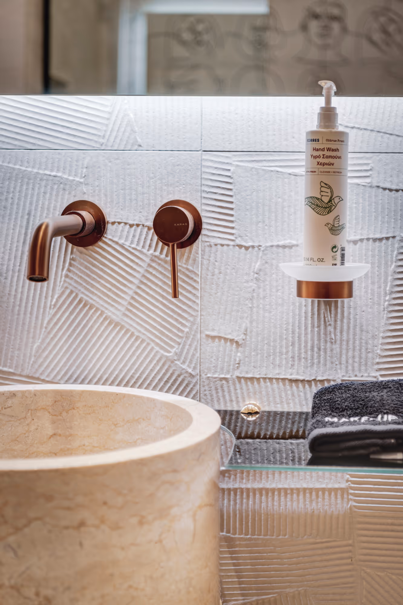 Modern bathroom sink with cylindrical stone basin, bronze faucet and handle, and a bottle of Korres hand wash on a glass shelf with a folded black towel.