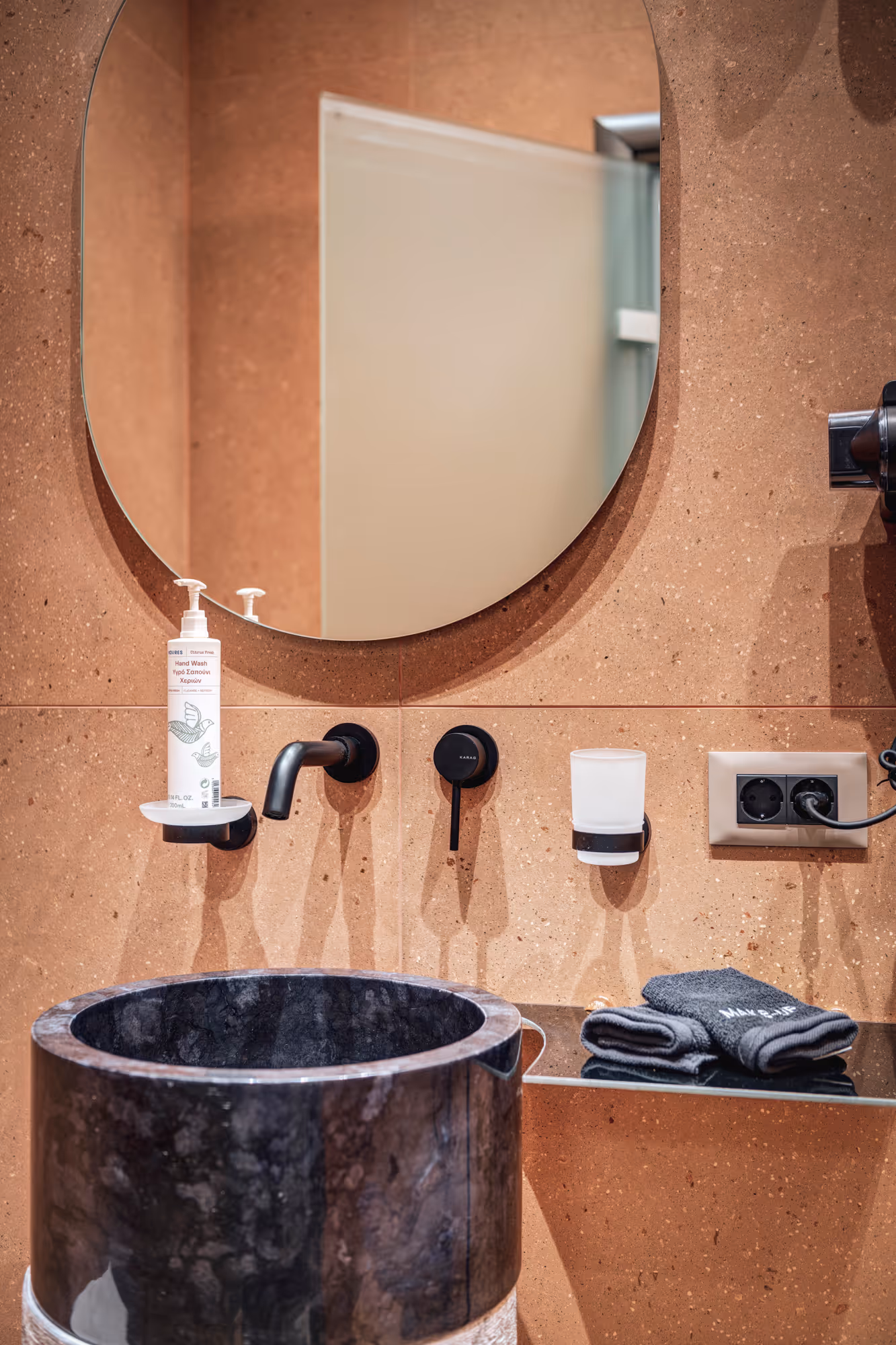 Modern bathroom sink area with a round mirror, black marble vessel sink, wall-mounted faucet, hand wash dispenser, and folded dark towels on a glass shelf.