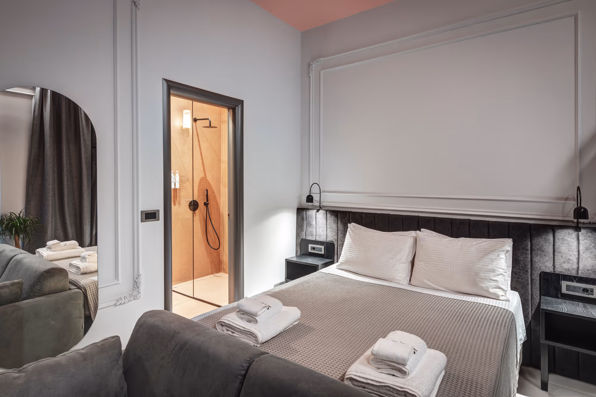 Modern hotel room with a double bed, folded towels, nightstands, a large oval mirror, and an open door leading to a shower area.