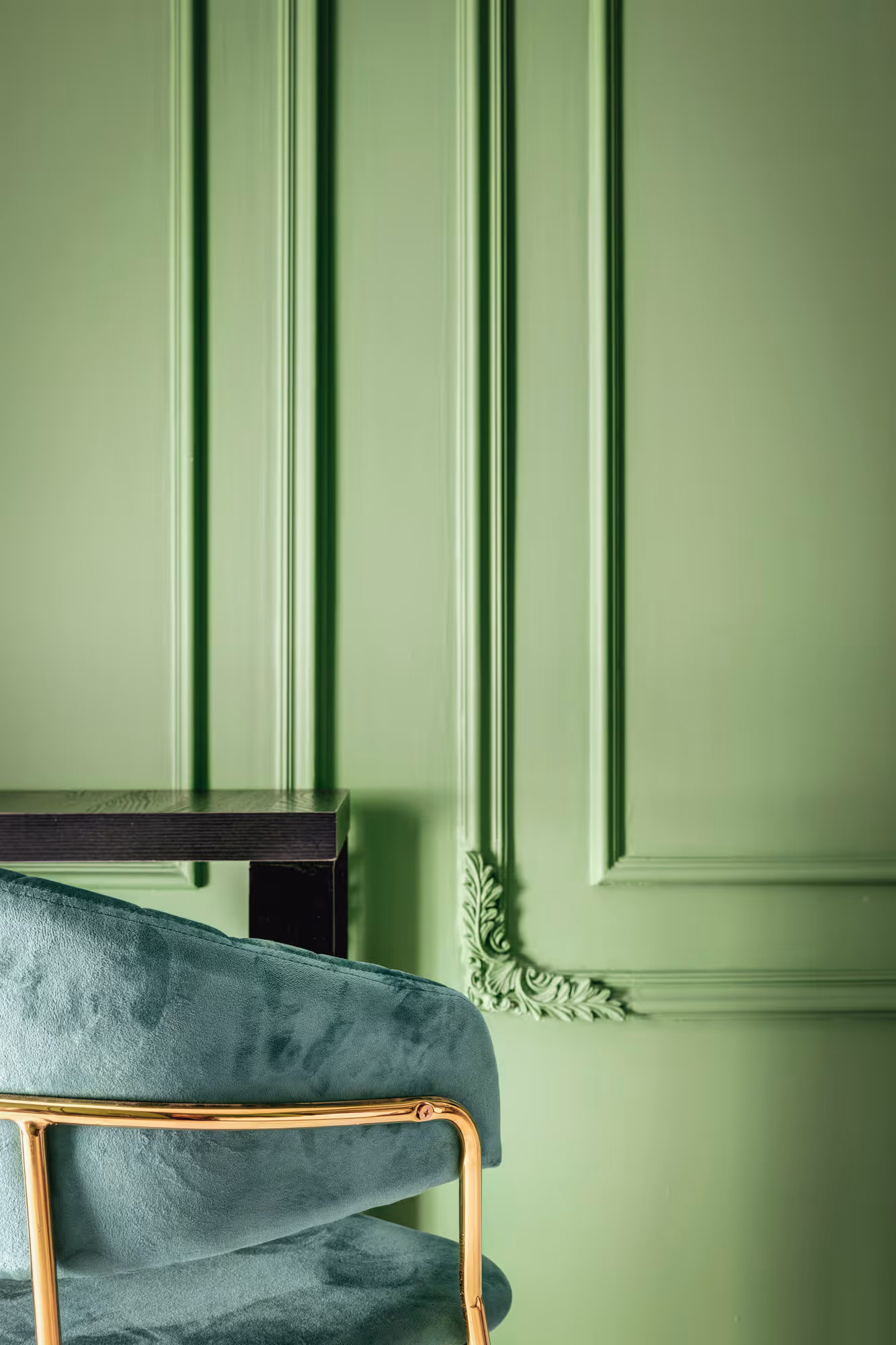 Close-up of a velvet green chair with gold metal frame next to a black table against a green wall with decorative molding.