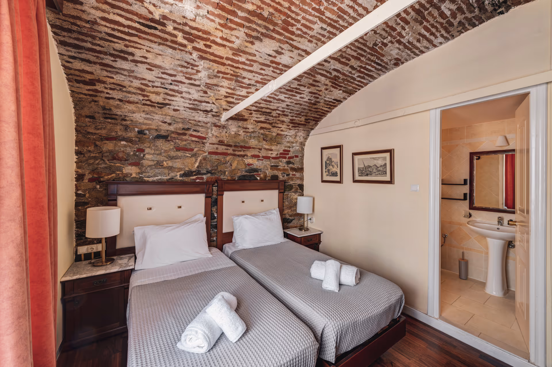 Hotel room with two single beds, rolled towels, bedside tables with lamps, exposed brick vaulted ceiling, and a bathroom with sink and mirror.