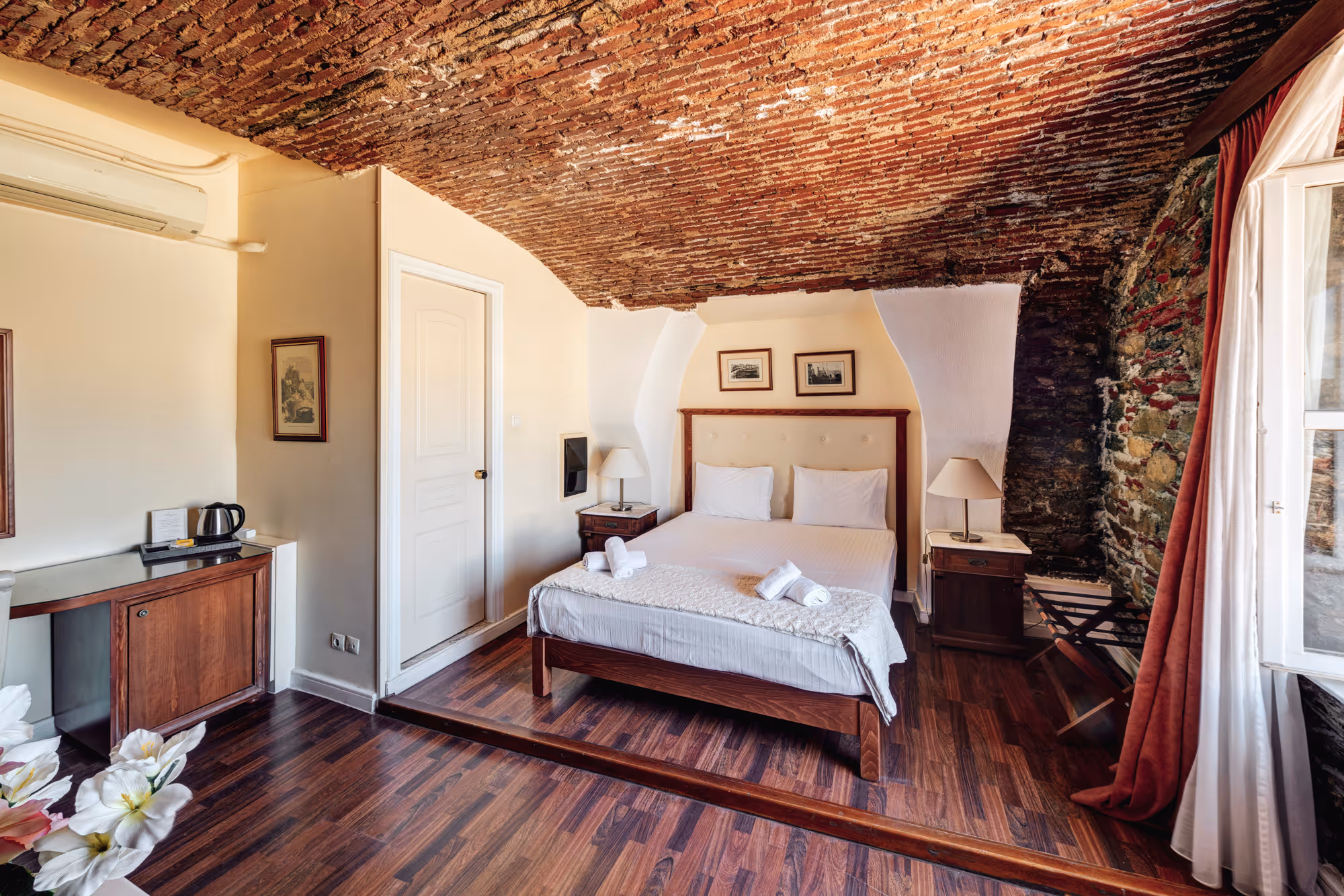 Cozy bedroom with a wooden bed, white bedding, two pillows, rolled towels on the bed, brick vaulted ceiling, wooden floor, and stone wall near window with red curtains.