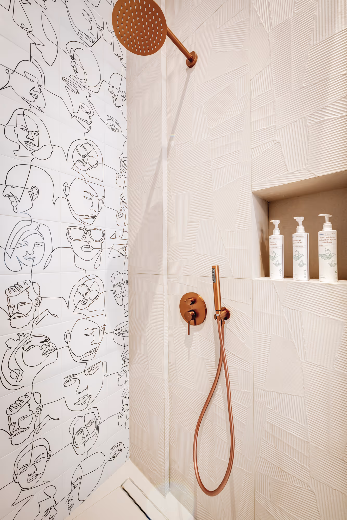 Modern shower area with white textured tiles, copper showerhead and handles, and wall art featuring black line-drawn faces.