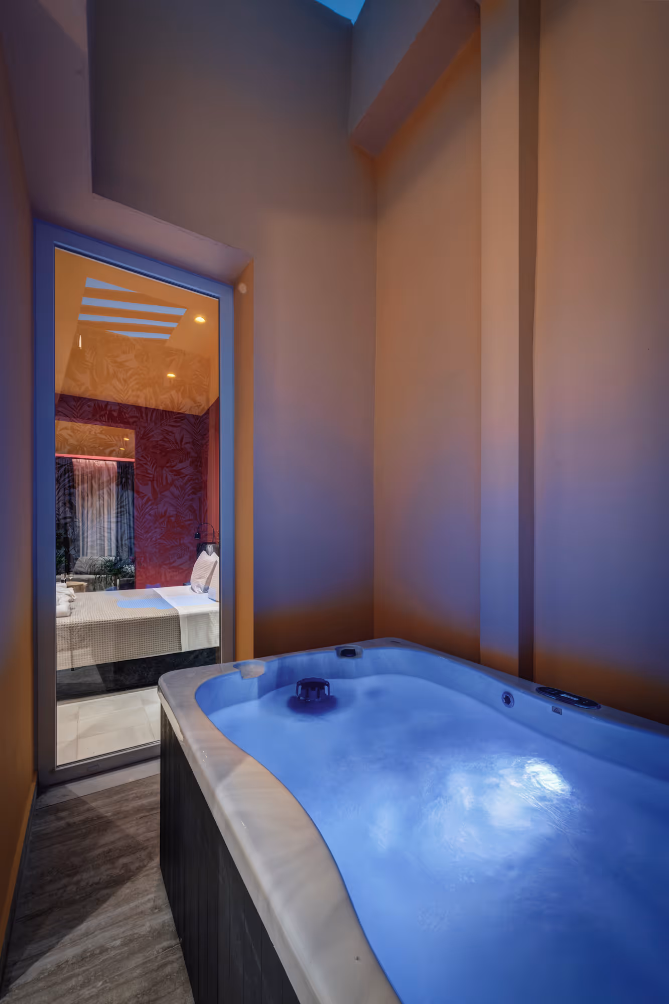 Blue-lit indoor hot tub next to a glass door showing a cozy bedroom with a bed and patterned wall.
