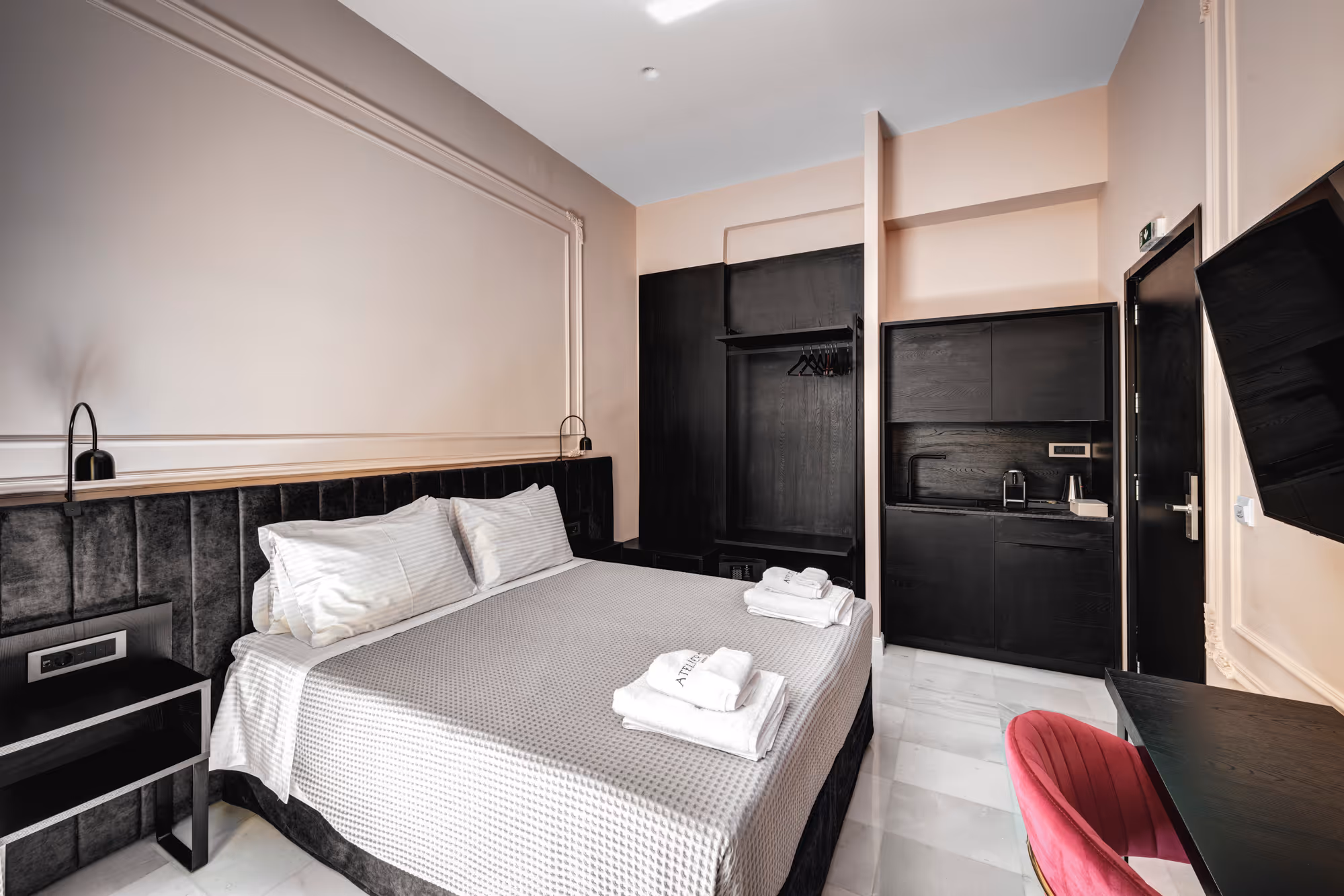 Modern hotel room with a large bed, black headboard, open black wardrobe, kitchenette, and a pink chair by a desk.
