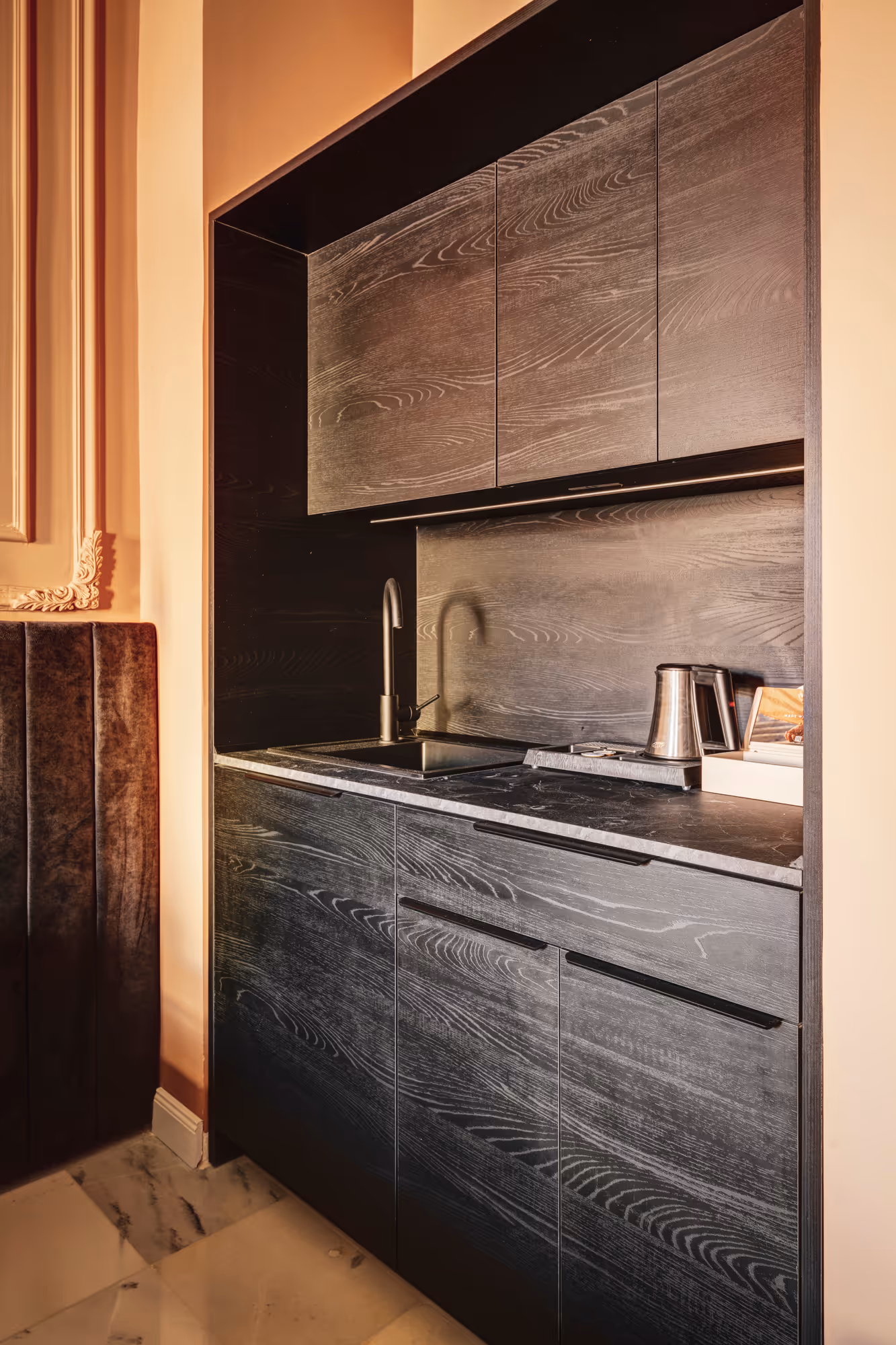 Compact modern kitchenette with dark wood cabinets, black countertop, stainless steel sink, and electric kettle.