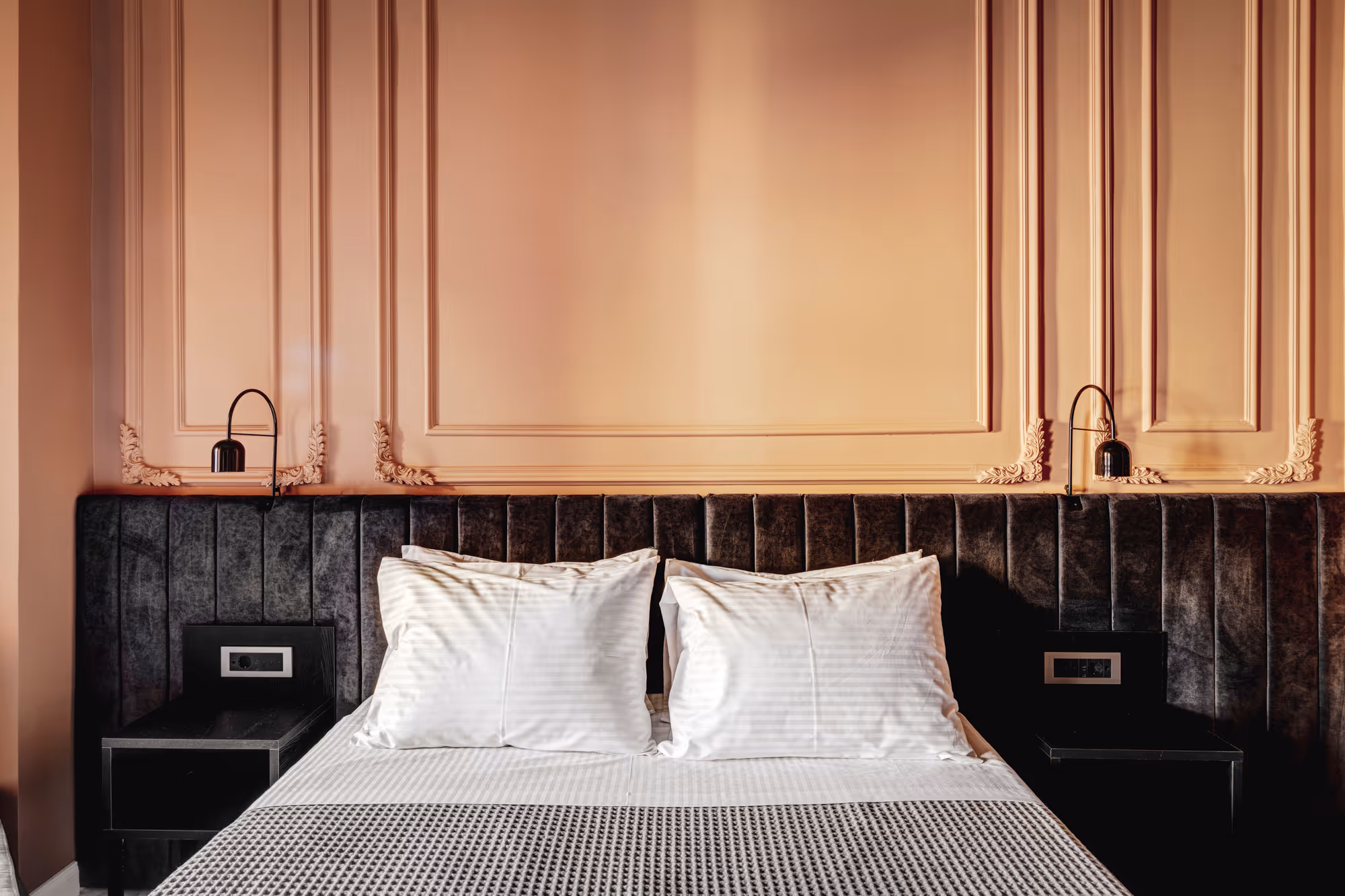 Modern bed with white pillows and a textured blanket against a dark velvet headboard and peach wall with decorative molding.