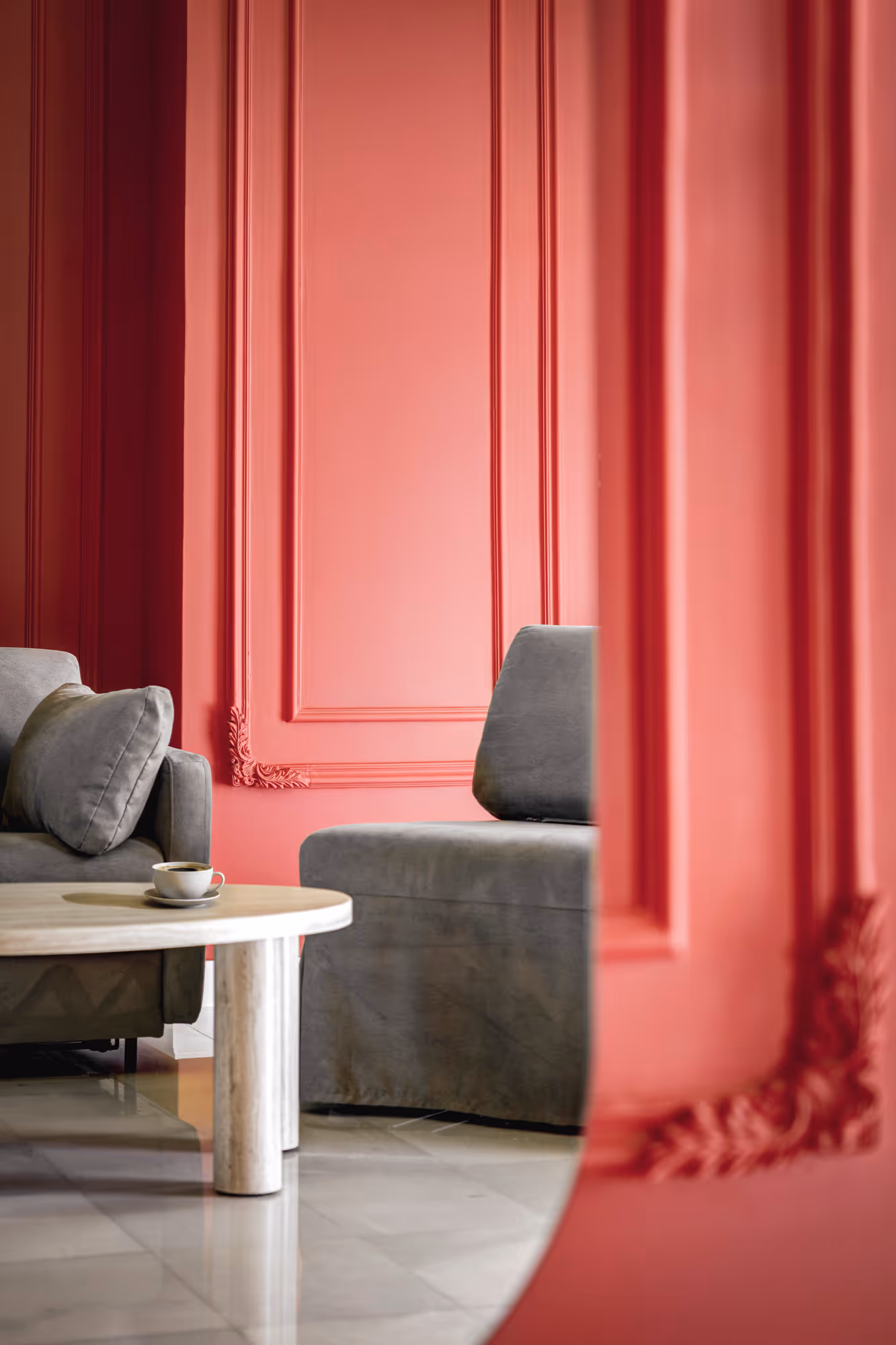 Gray velvet sofa and cushioned chair in a room with coral-pink decorative wall paneling and a round white marble coffee table with a cup of coffee.