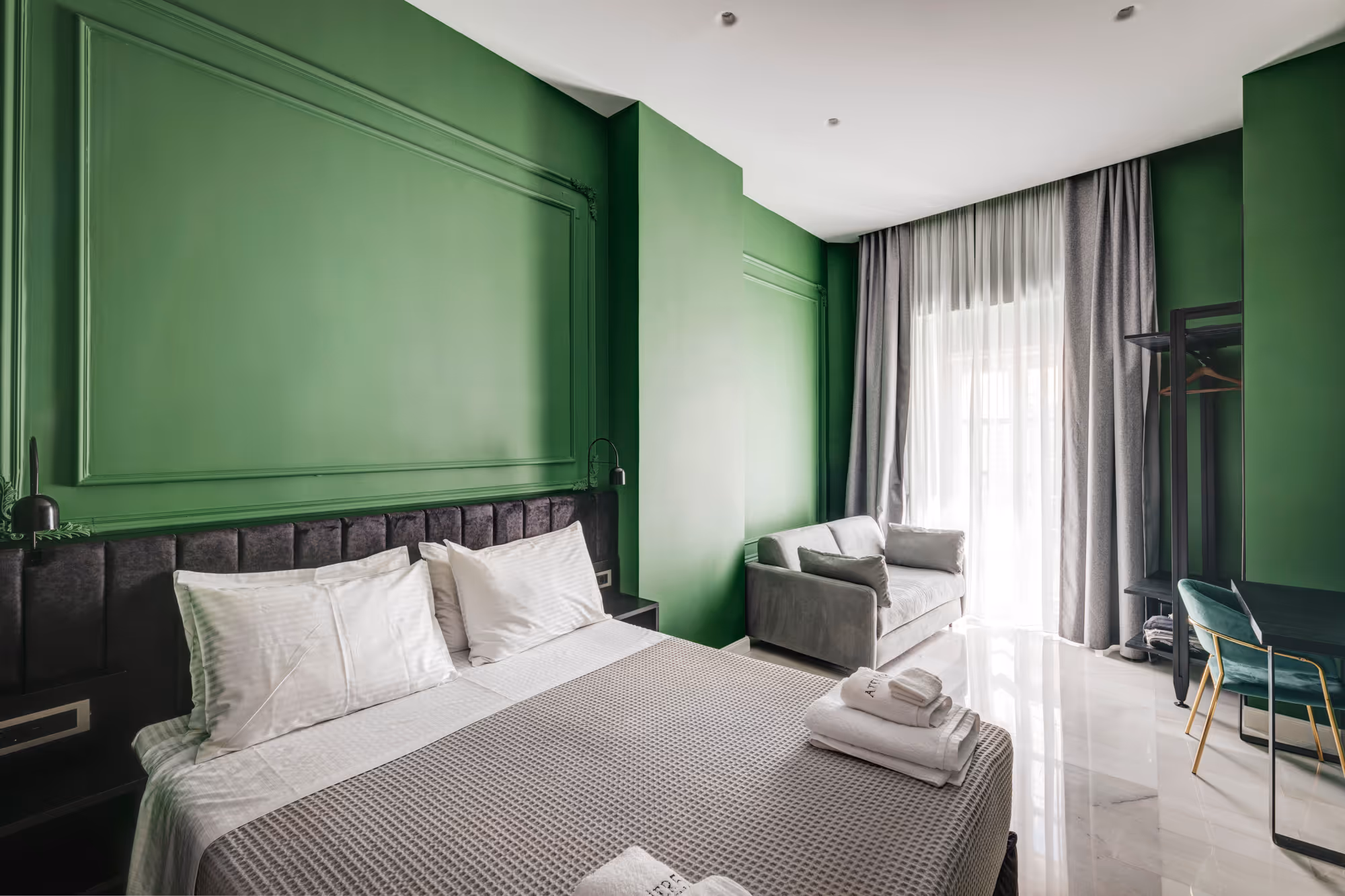 Modern hotel room with vibrant green walls, a large bed with white pillows, a gray sofa, and a desk with a chair near sheer curtains.