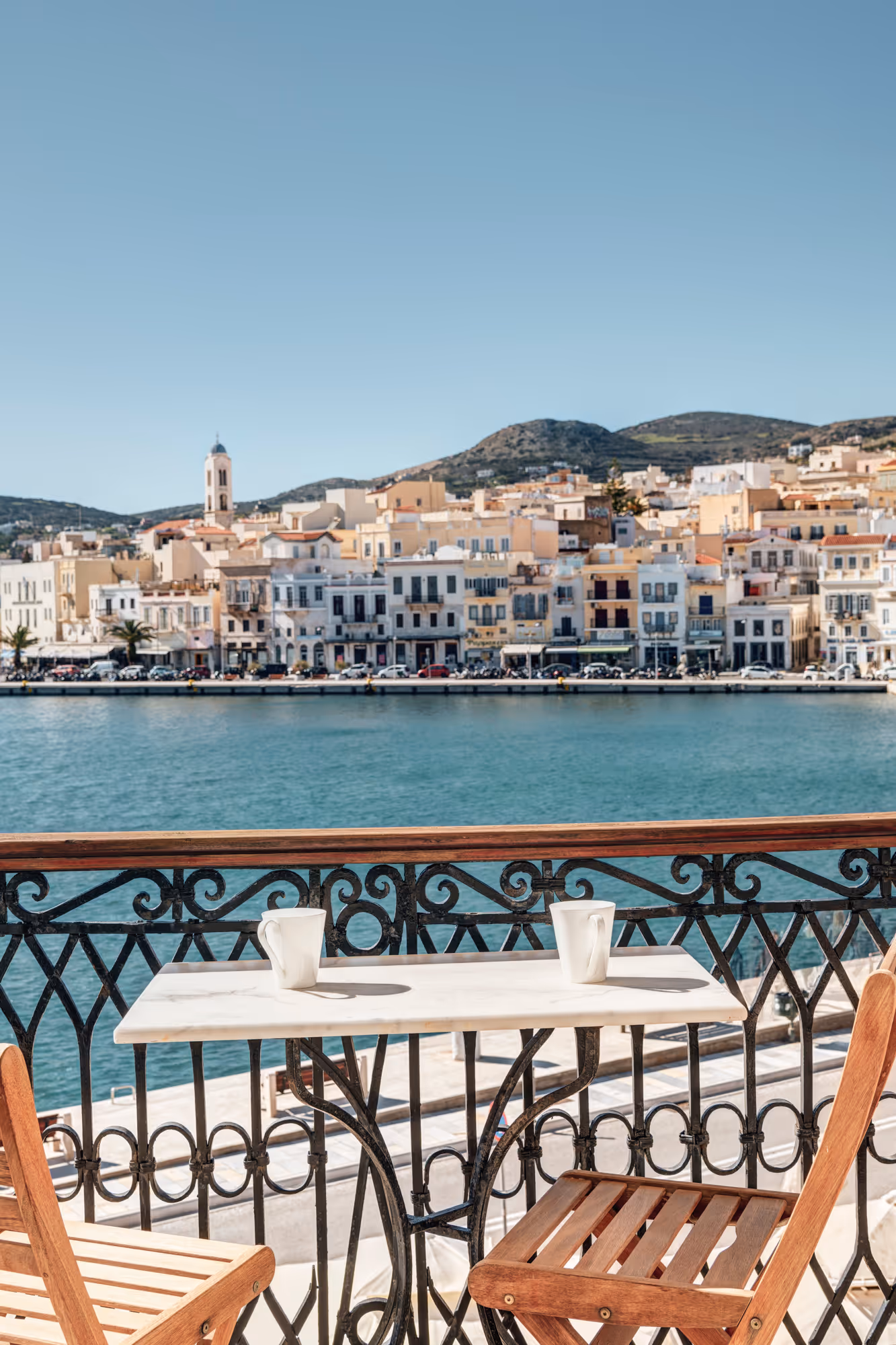 Two wooden chairs and a small table with cups on a balcony overlooking a waterfront town with buildings and hills in the background.