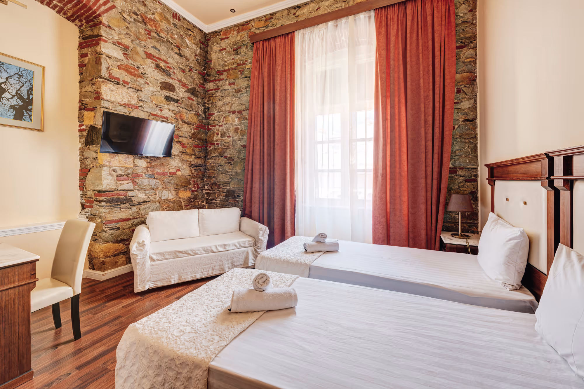 Hotel room with twin beds, white linens, rolled towels, a white sofa, stone accent wall, and a window with sheer and red curtains.