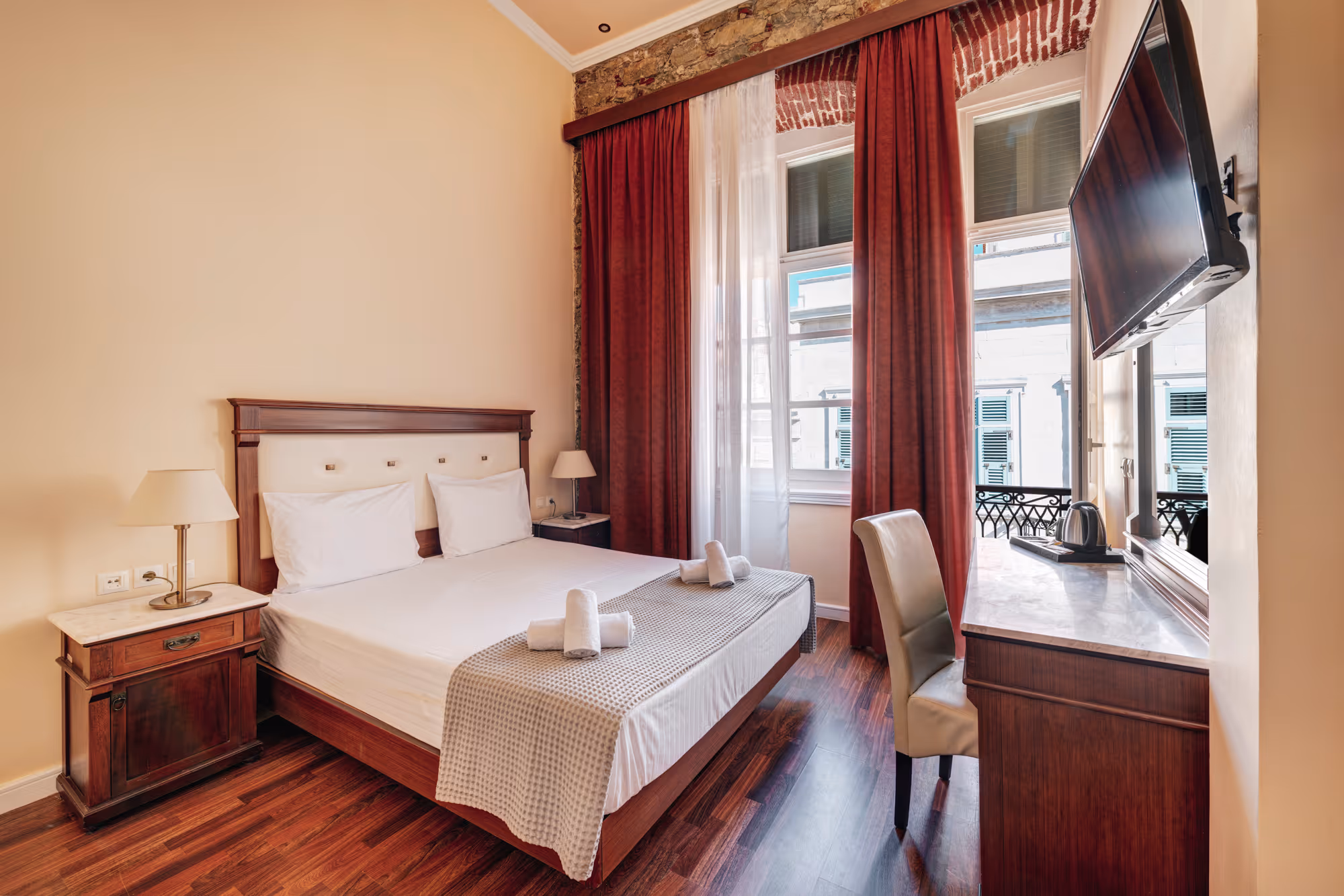 Neat hotel room with double bed, white linens, brown wooden furniture, red curtains, and a wall-mounted TV.
