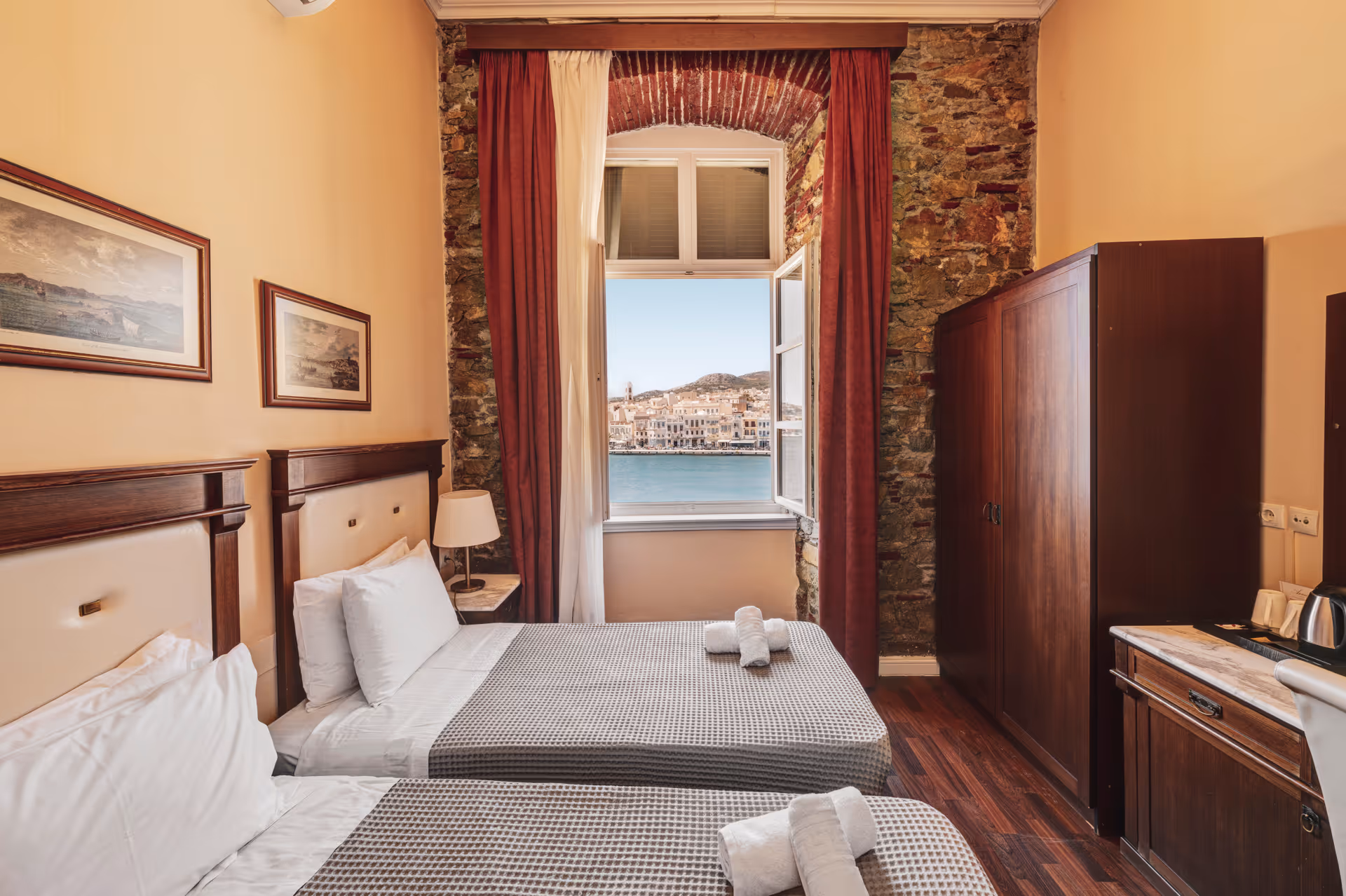 Cozy hotel room with two twin beds, wooden furniture, exposed brick window framing, and a view of a waterfront town through an open window.
