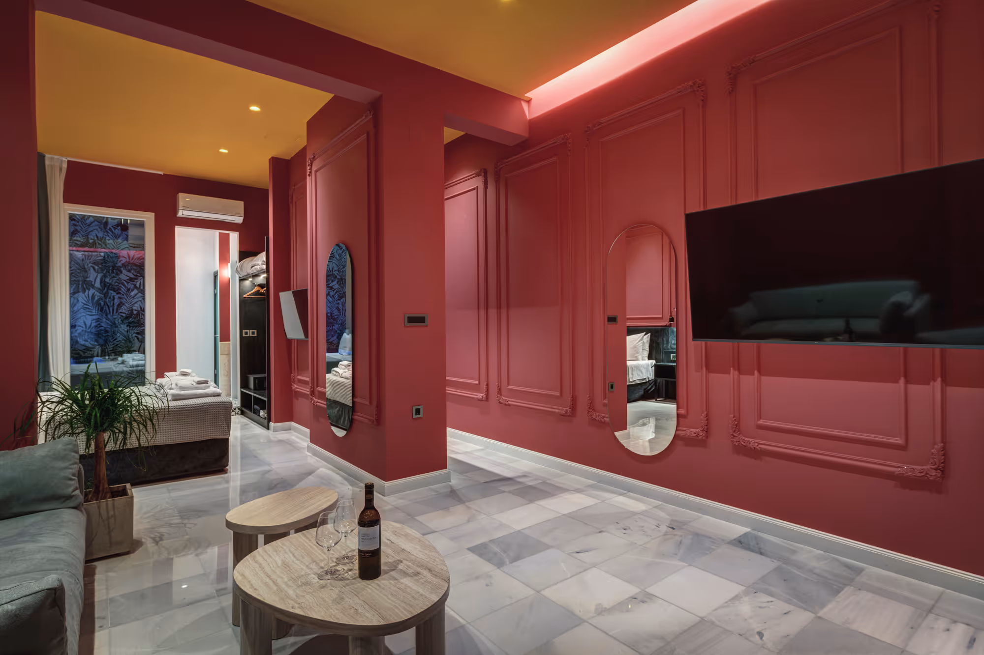 Modern hotel room with red paneled walls, yellow ceiling, marble floor, a sofa, coffee tables with a wine bottle and glasses, and a mounted flat-screen TV.