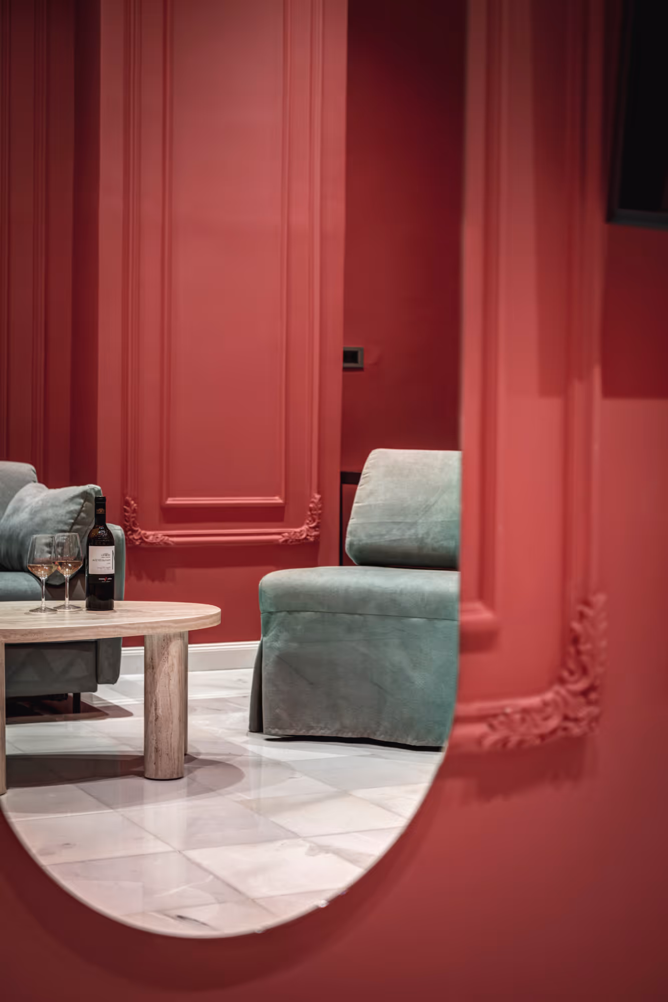 Reflection in a mirror showing a room with red walls, a wooden coffee table with a wine bottle and two glasses, and green upholstered chairs.
