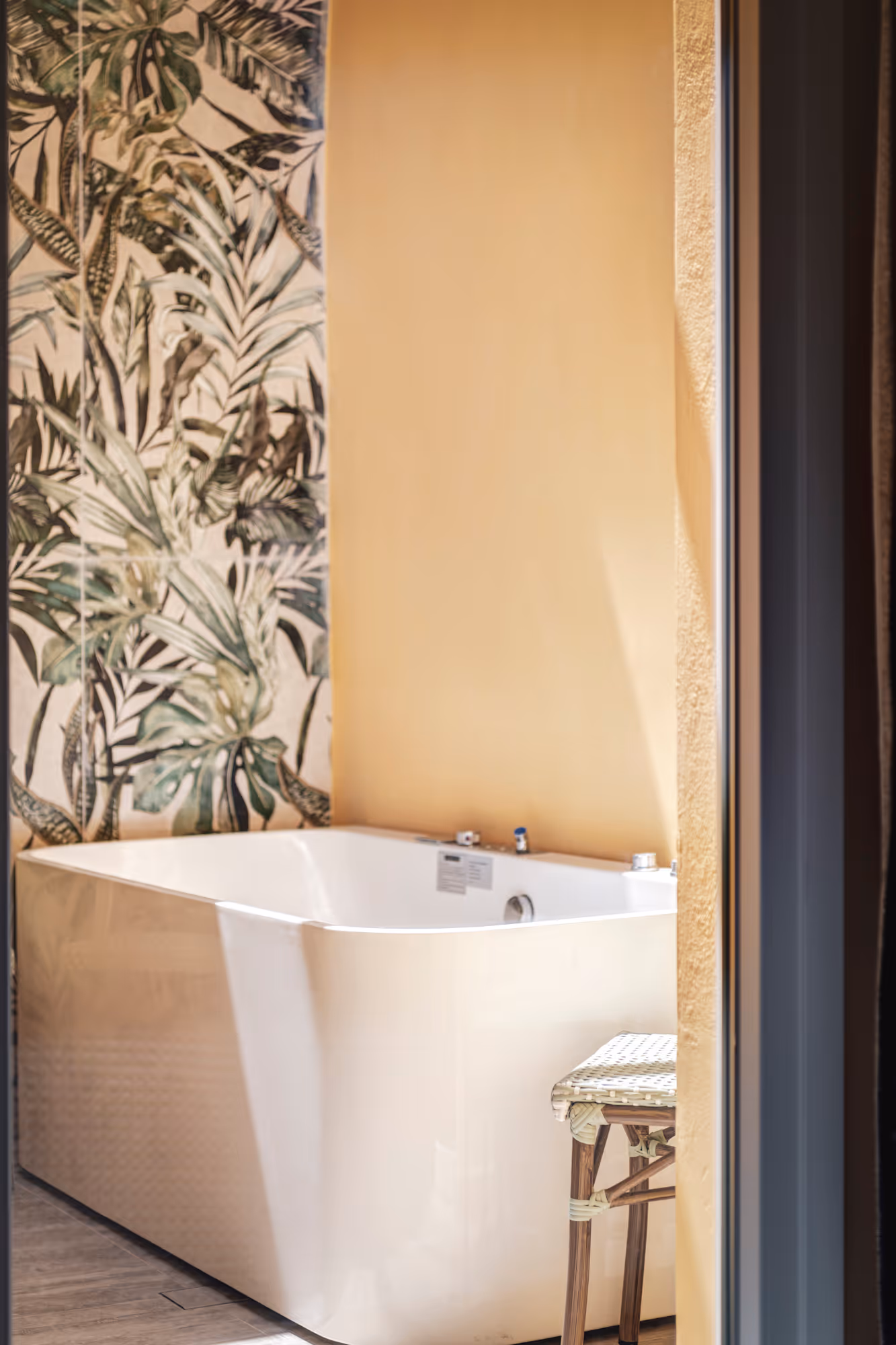 White bathtub next to a wall with tropical leaf patterned tiles and a wooden stool, in a sunlit bathroom.