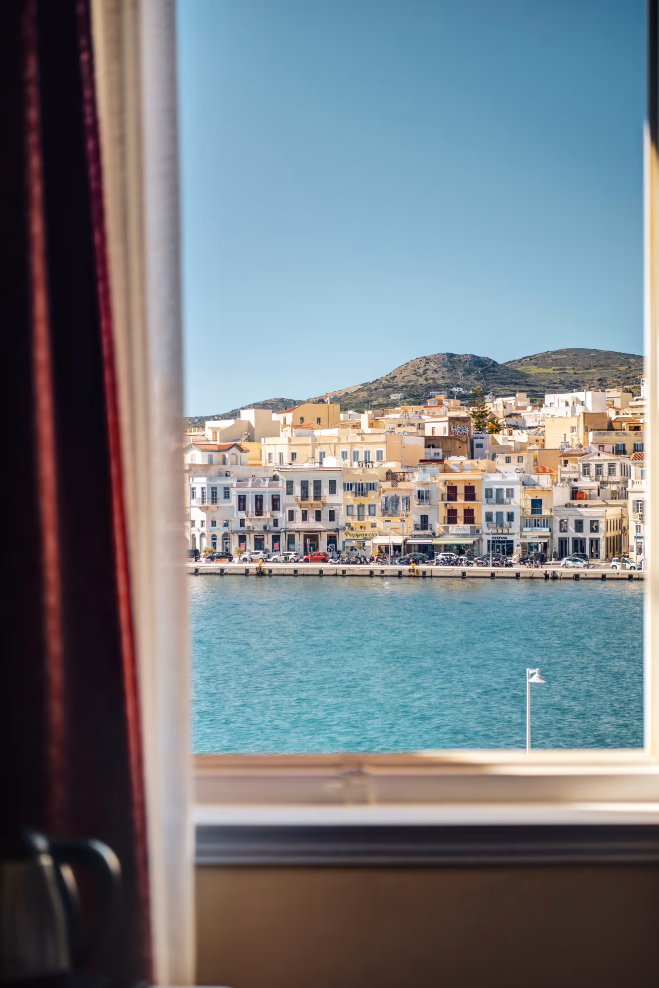 View through a window showing a waterfront town with buildings, turquoise water, and hills in the background under a clear blue sky.