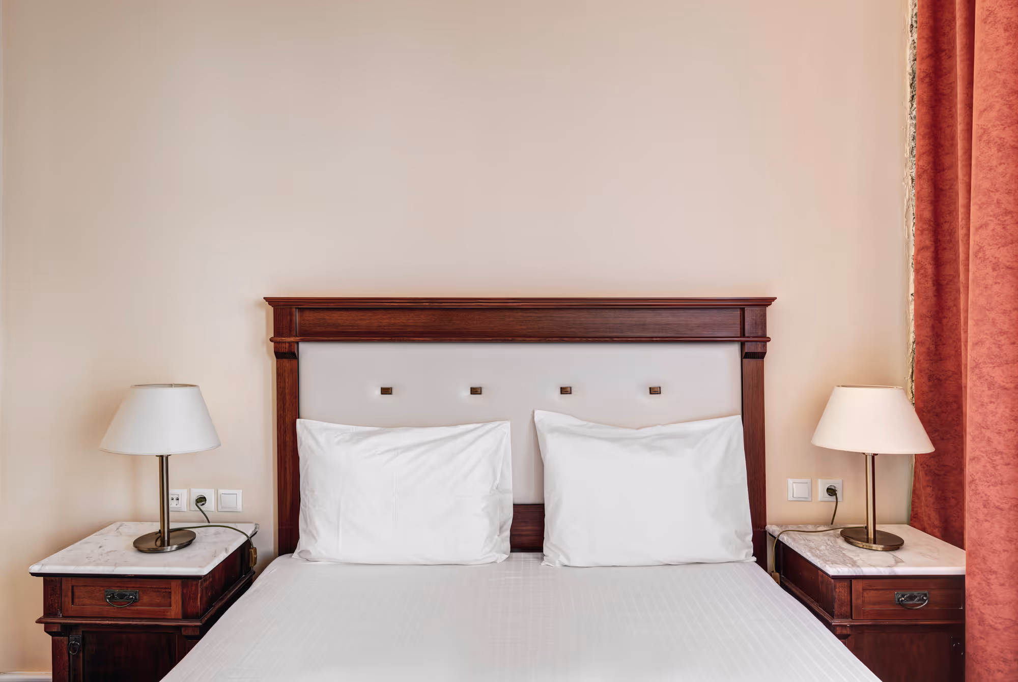 Neatly made bed with white pillows and sheets, dark wood headboard, flanked by two nightstands with marble tops and table lamps.