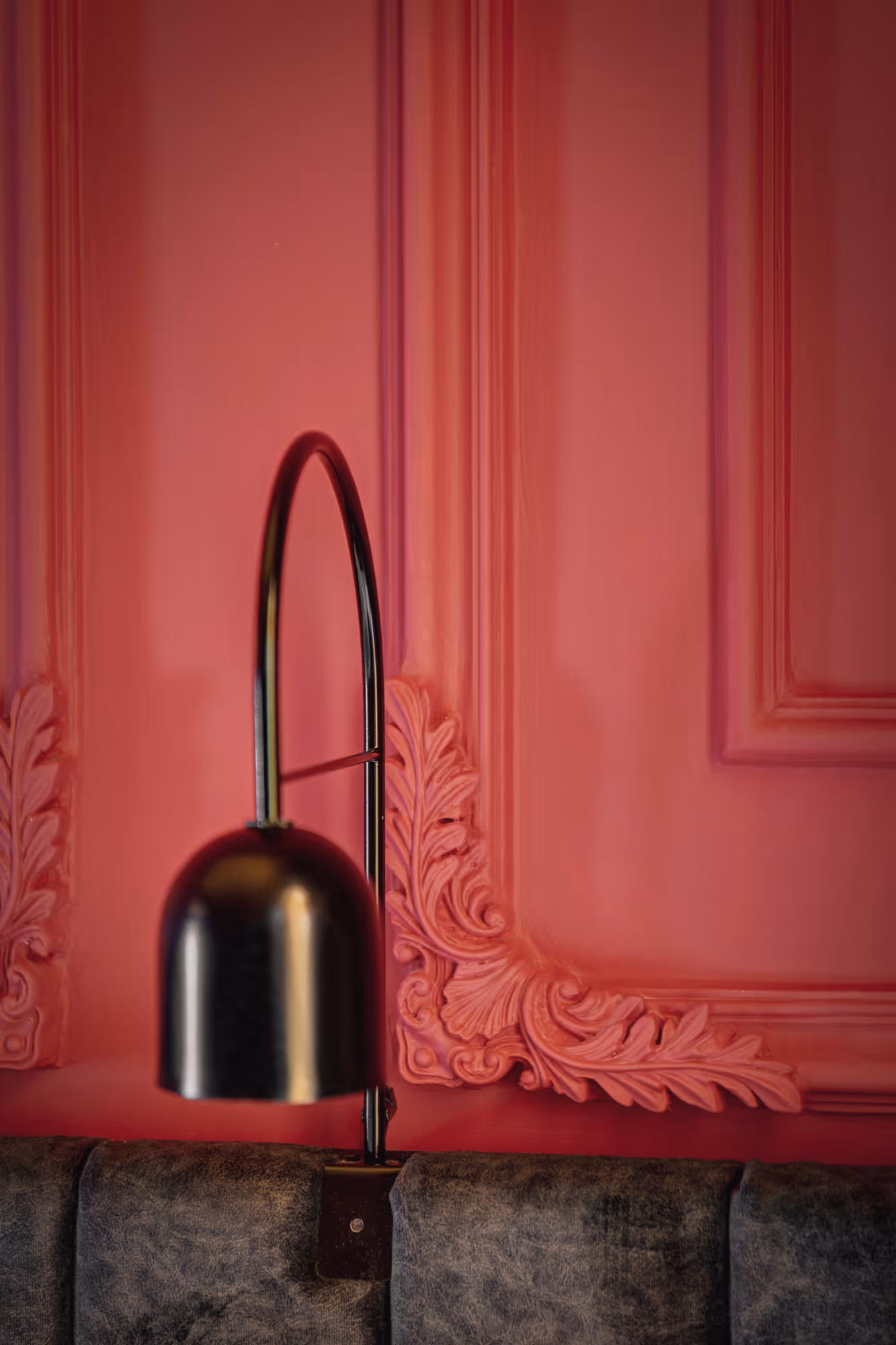 Black curved wall lamp mounted above a dark upholstered bench against a pink wall with decorative molding.