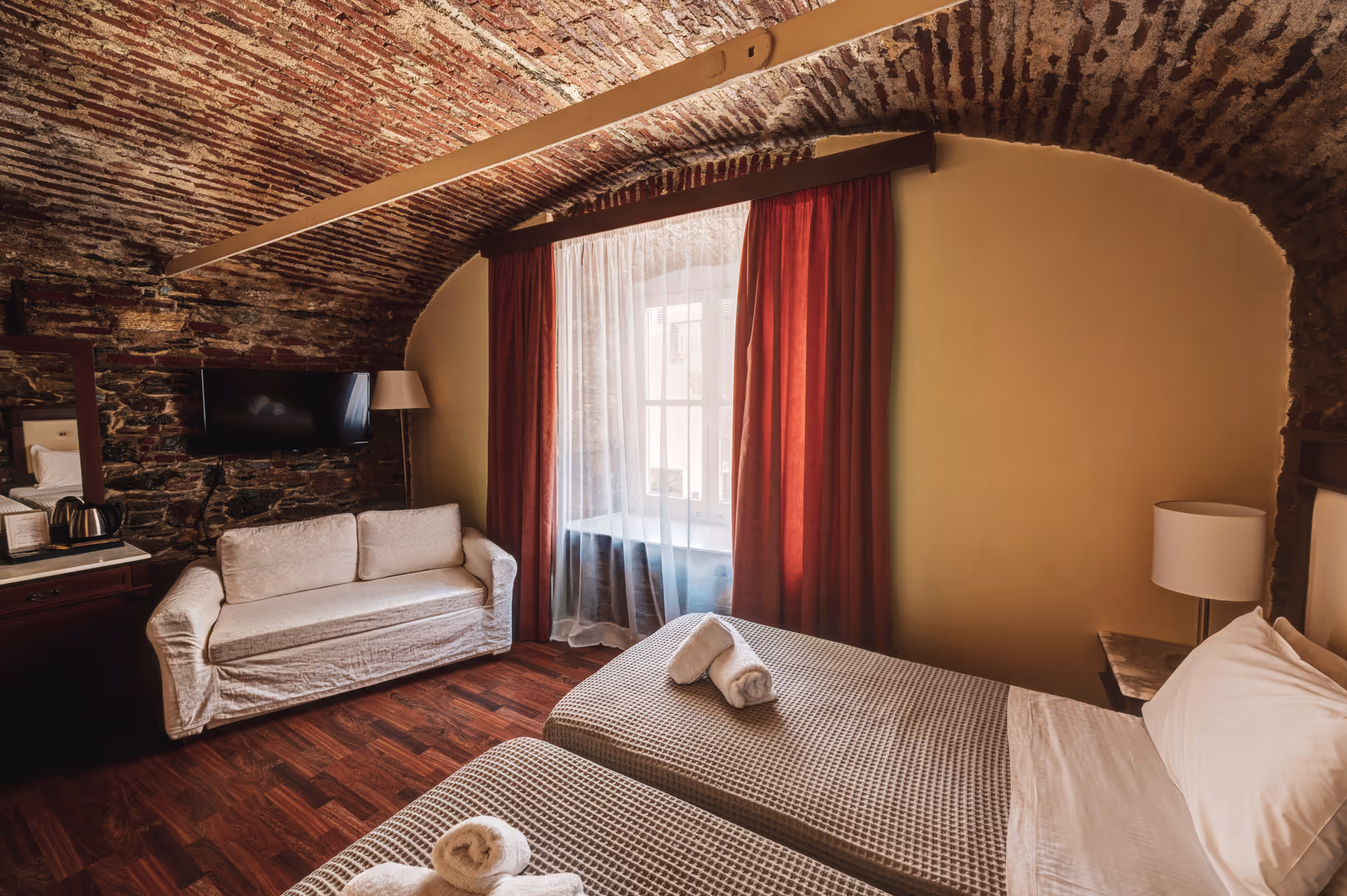 Hotel room with twin beds topped with folded towels, a white couch, exposed brick vaulted ceiling, and a window with red curtains.
