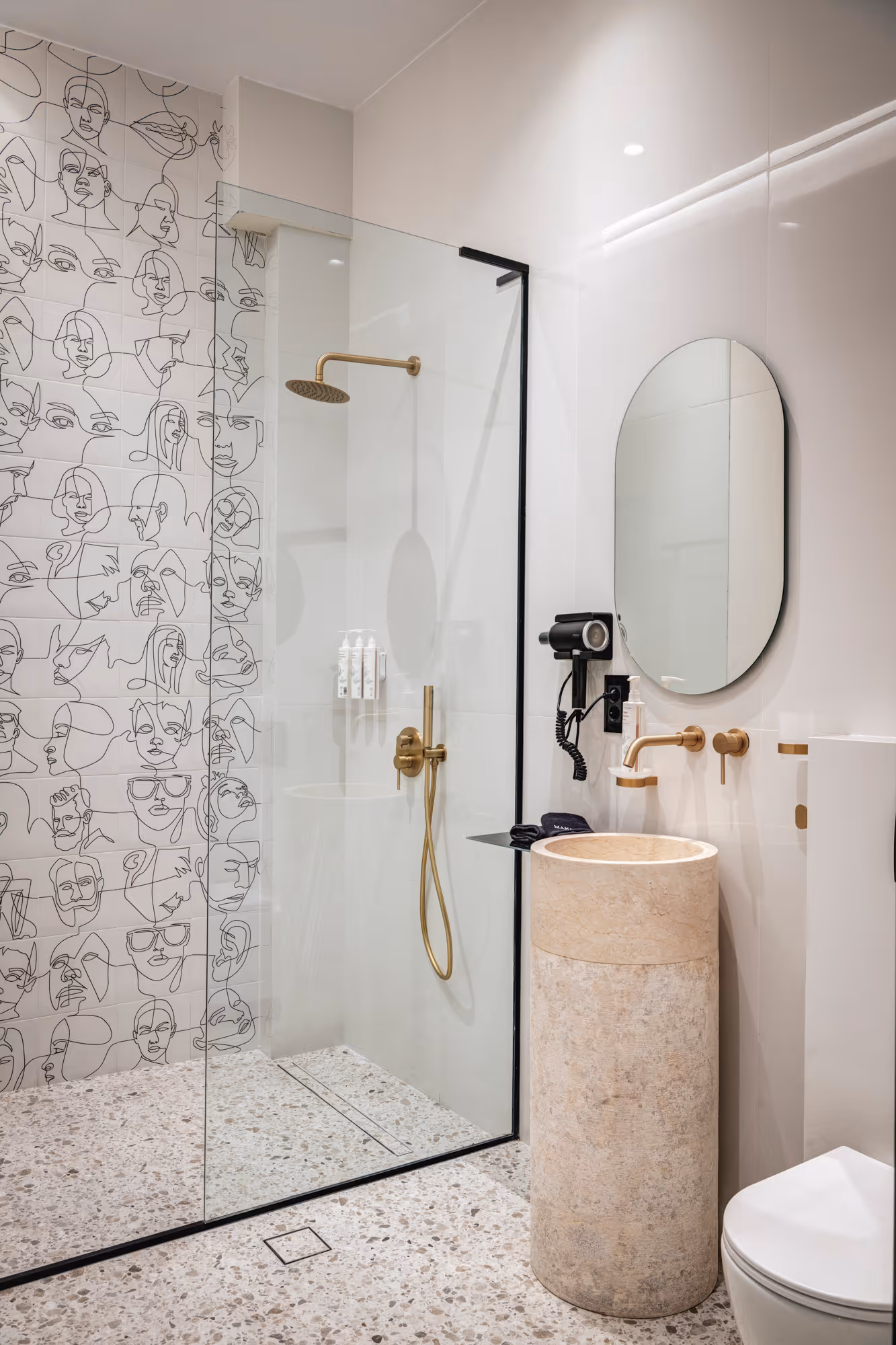 Modern bathroom with terrazzo floor, walk-in shower with gold fixtures, wall with black line art faces, round mirror, cylindrical stone sink, and white toilet.