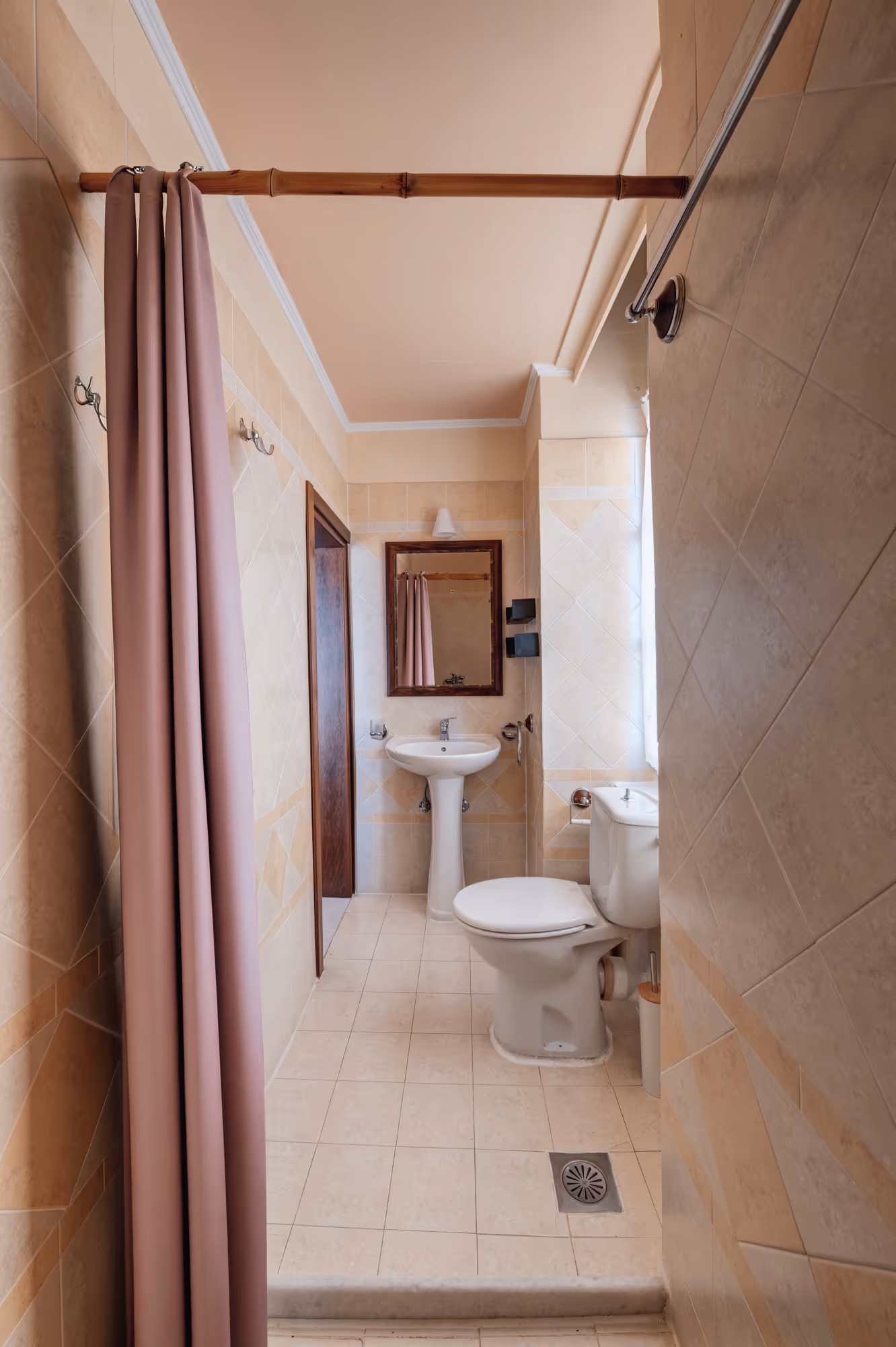Small beige tiled bathroom with white pedestal sink, toilet, mirror, and mauve shower curtain.