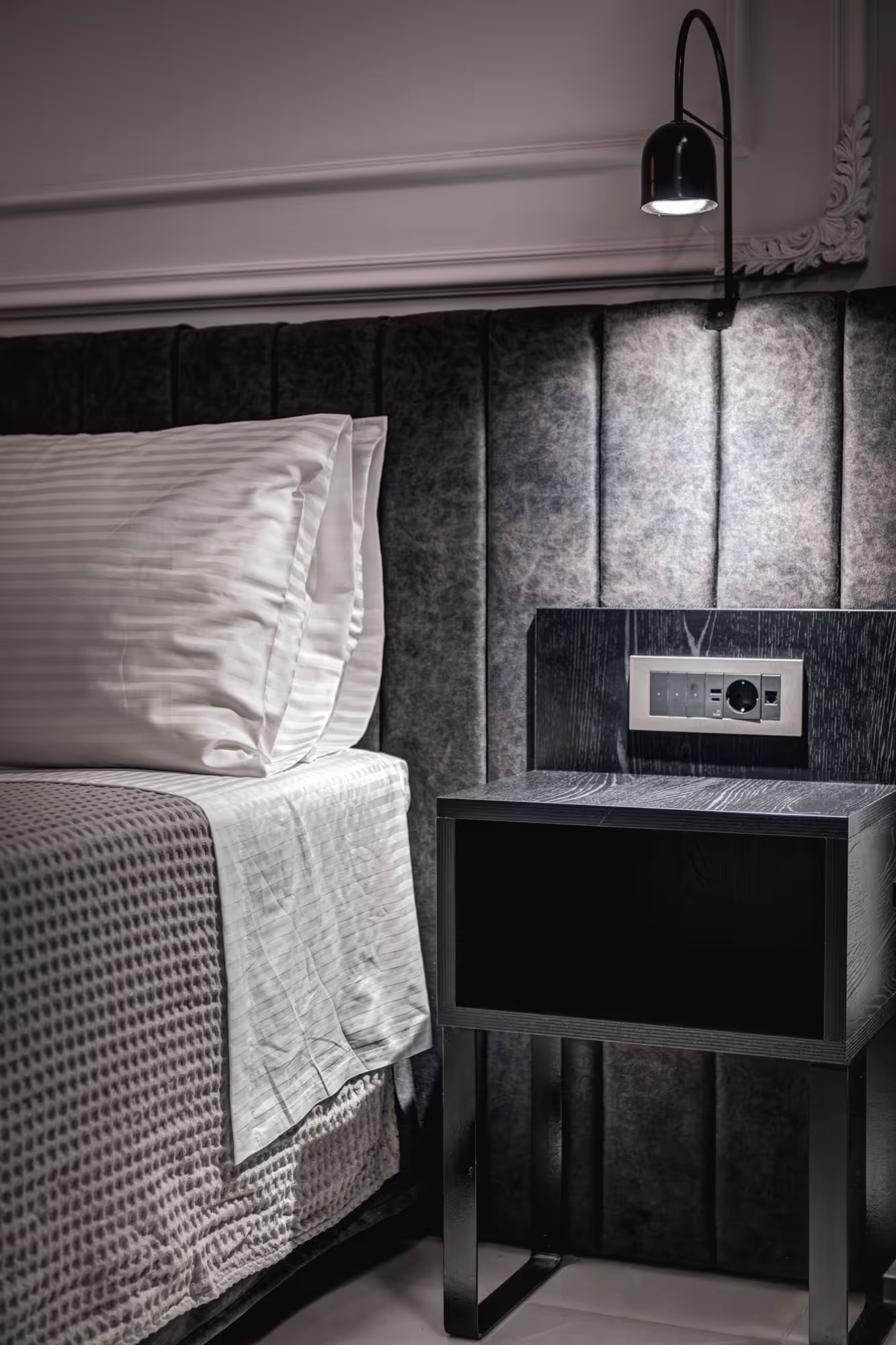 Close-up of a bed with white striped pillows and blanket next to a black bedside table with built-in power outlets and a wall lamp above.