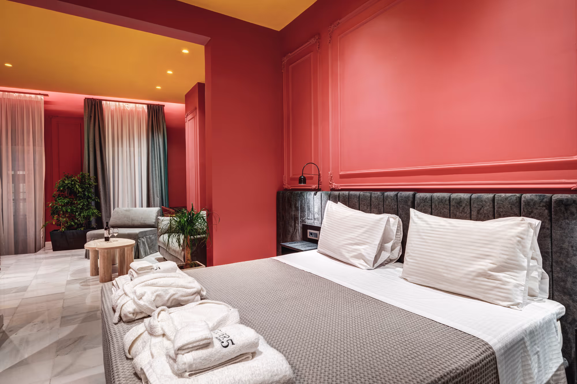 Modern hotel room with a large bed, gray headboard, red walls, yellow ceiling, armchair, and white bathrobes on the bed.