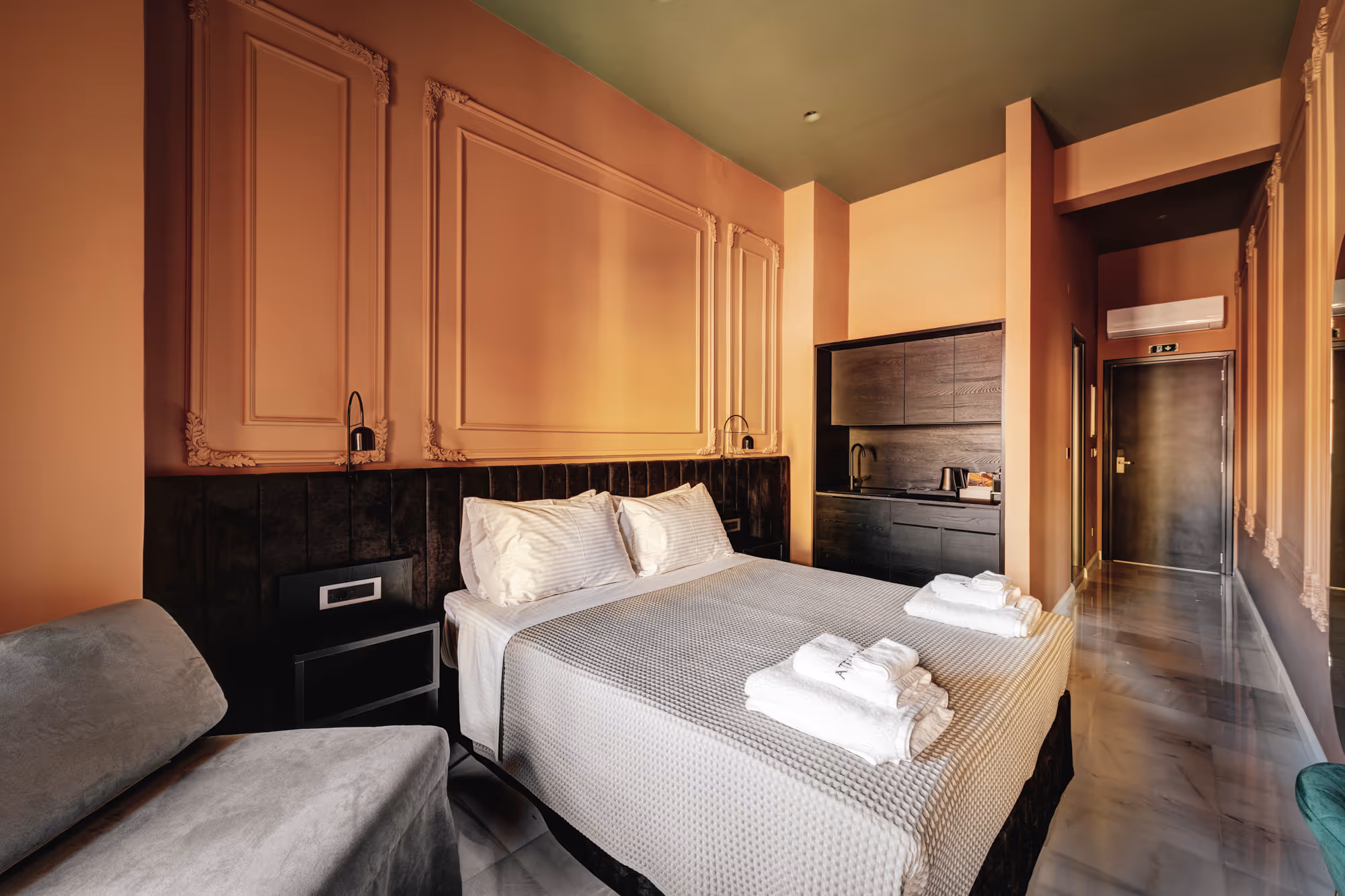 Modern hotel room with a double bed, white linens, folded towels on the bed, a gray armchair, peach walls with decorative molding, and a kitchenette area.