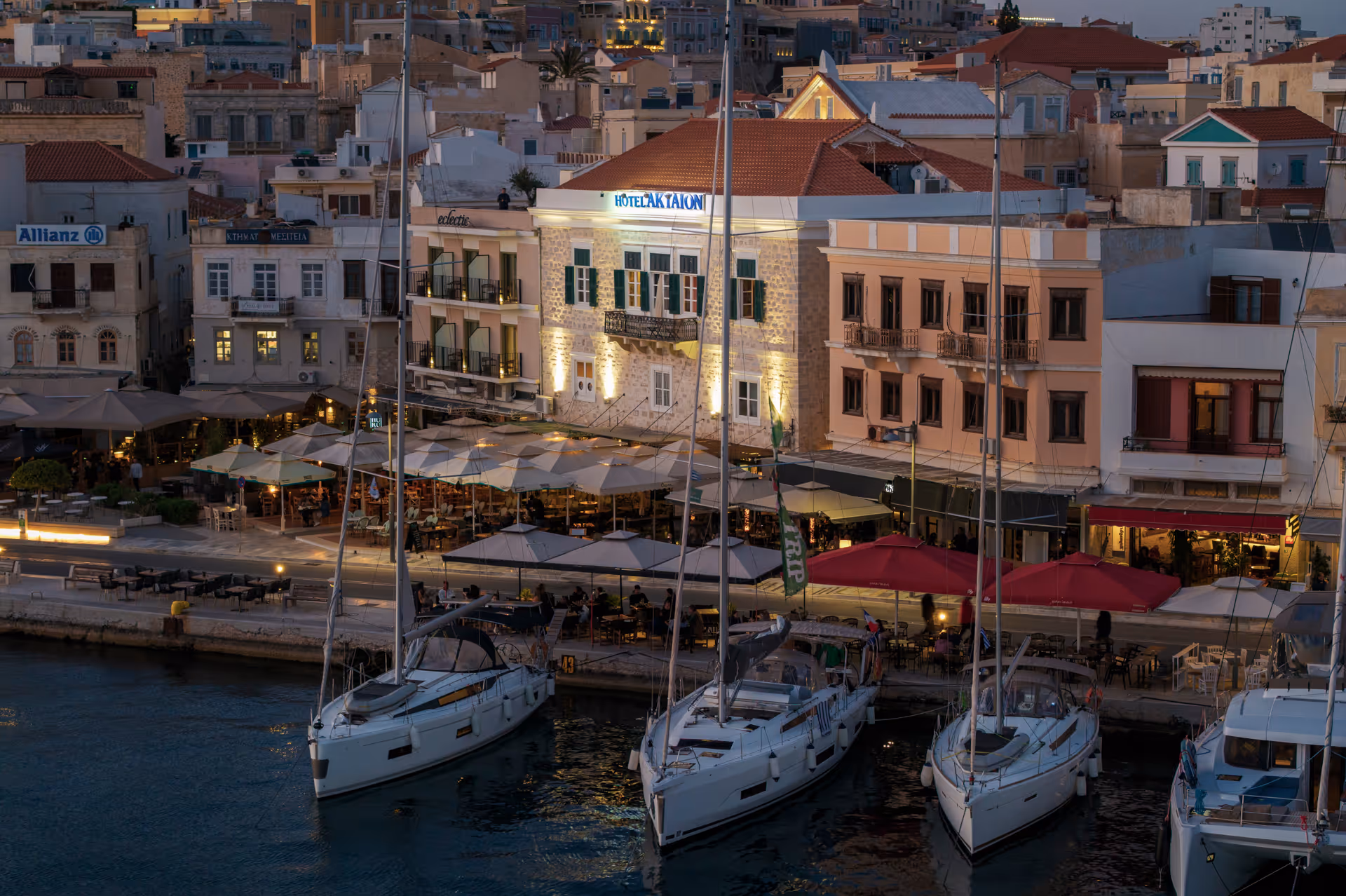 Evening view of a harbor with sailboats docked and waterfront buildings with outdoor seating and umbrellas.