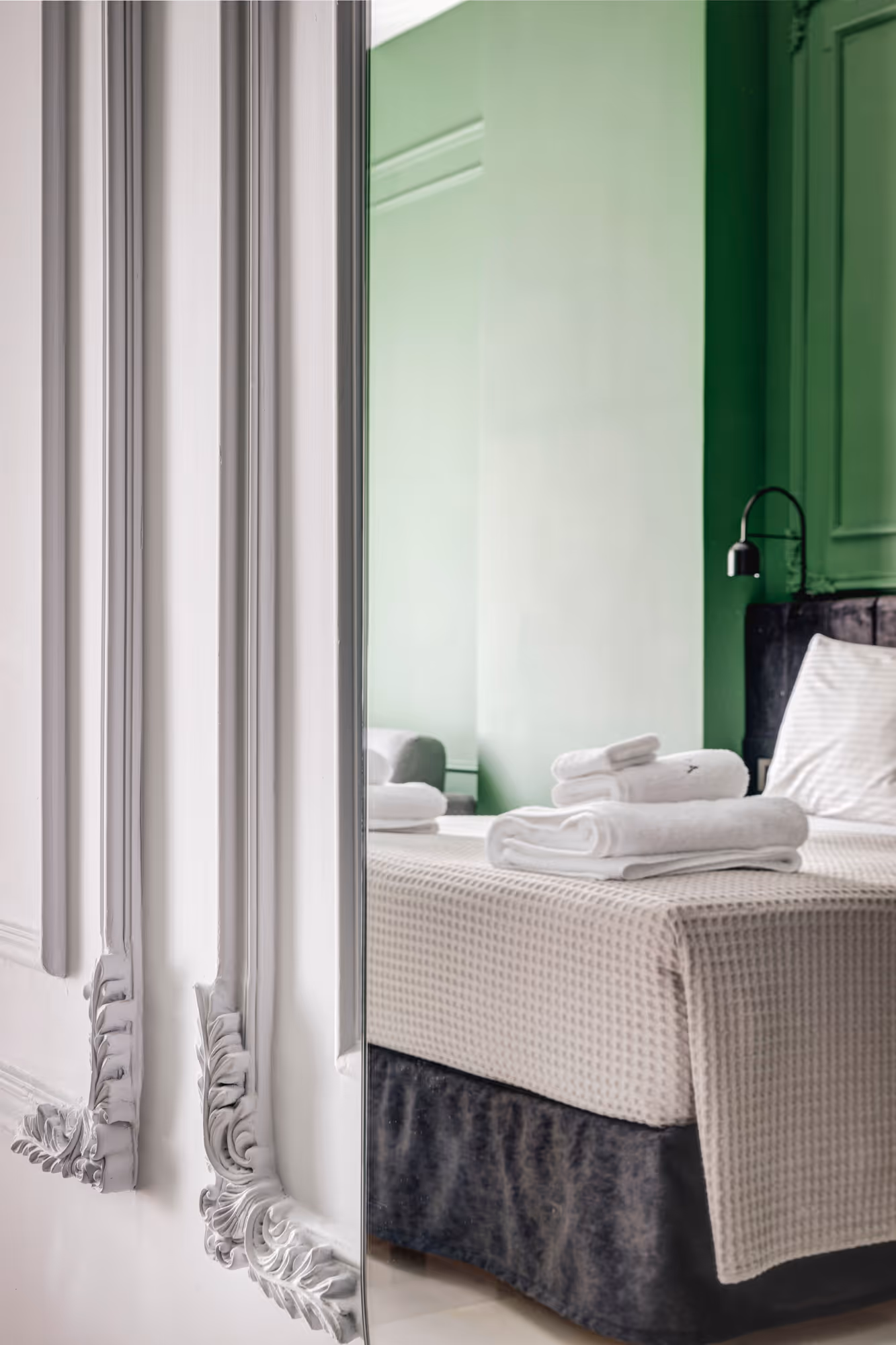 Modern bedroom reflected in a decorative mirror with a neatly made bed covered with beige textured bedding and folded white towels on top against a green wall.