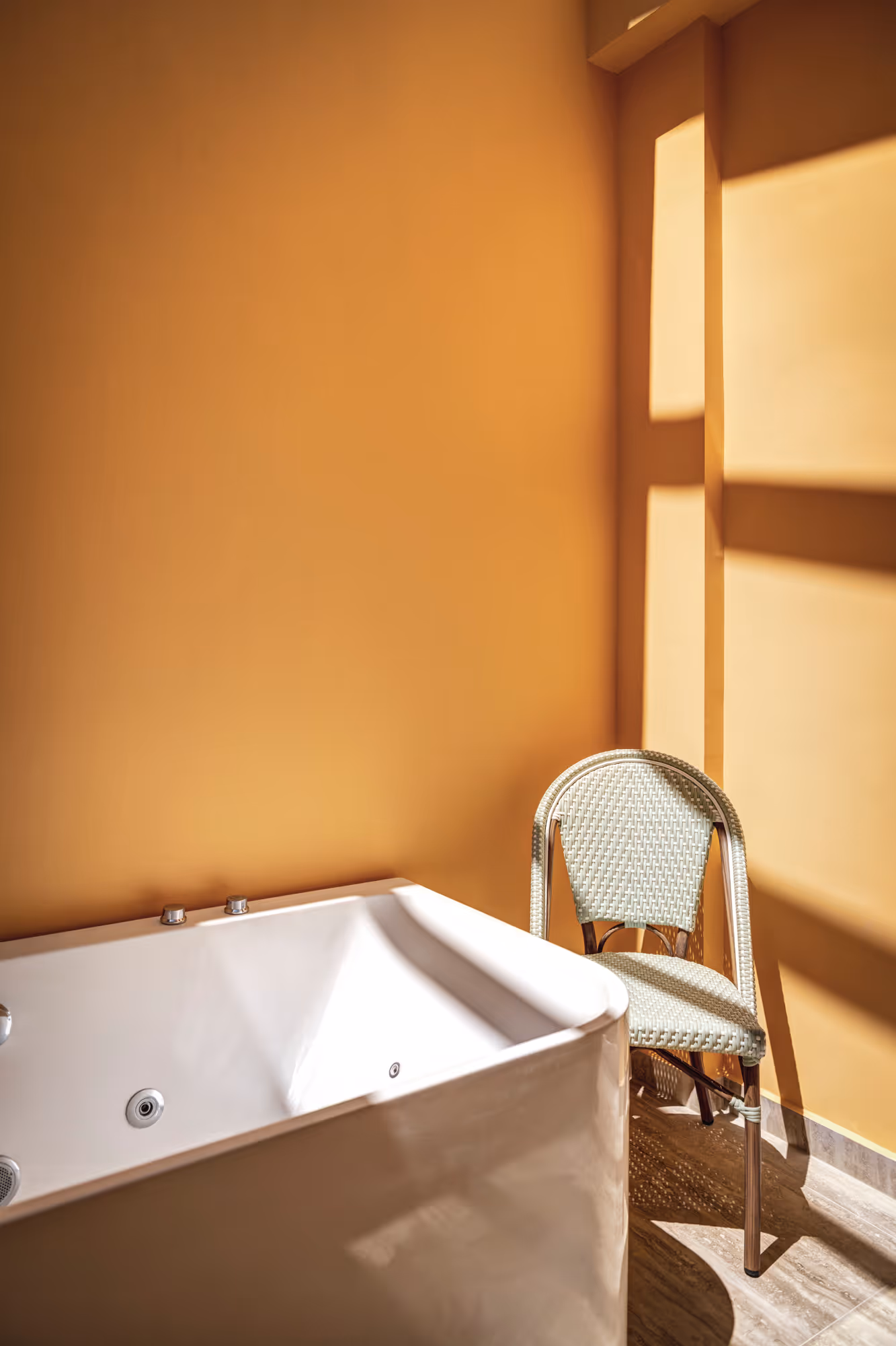 White jacuzzi bathtub next to a woven chair against a mustard yellow wall with sunlight creating shadows.