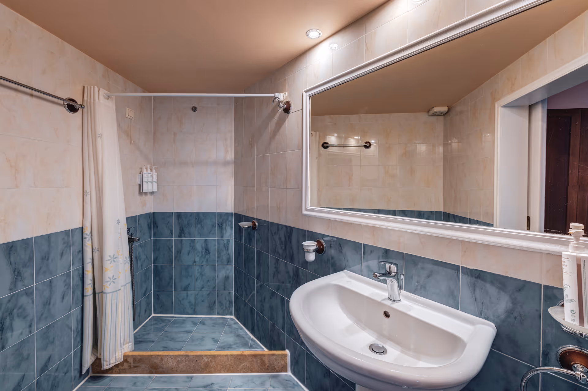 Modern bathroom with blue and beige tiled walls, a white pedestal sink, large framed mirror, and a shower area with a white curtain.