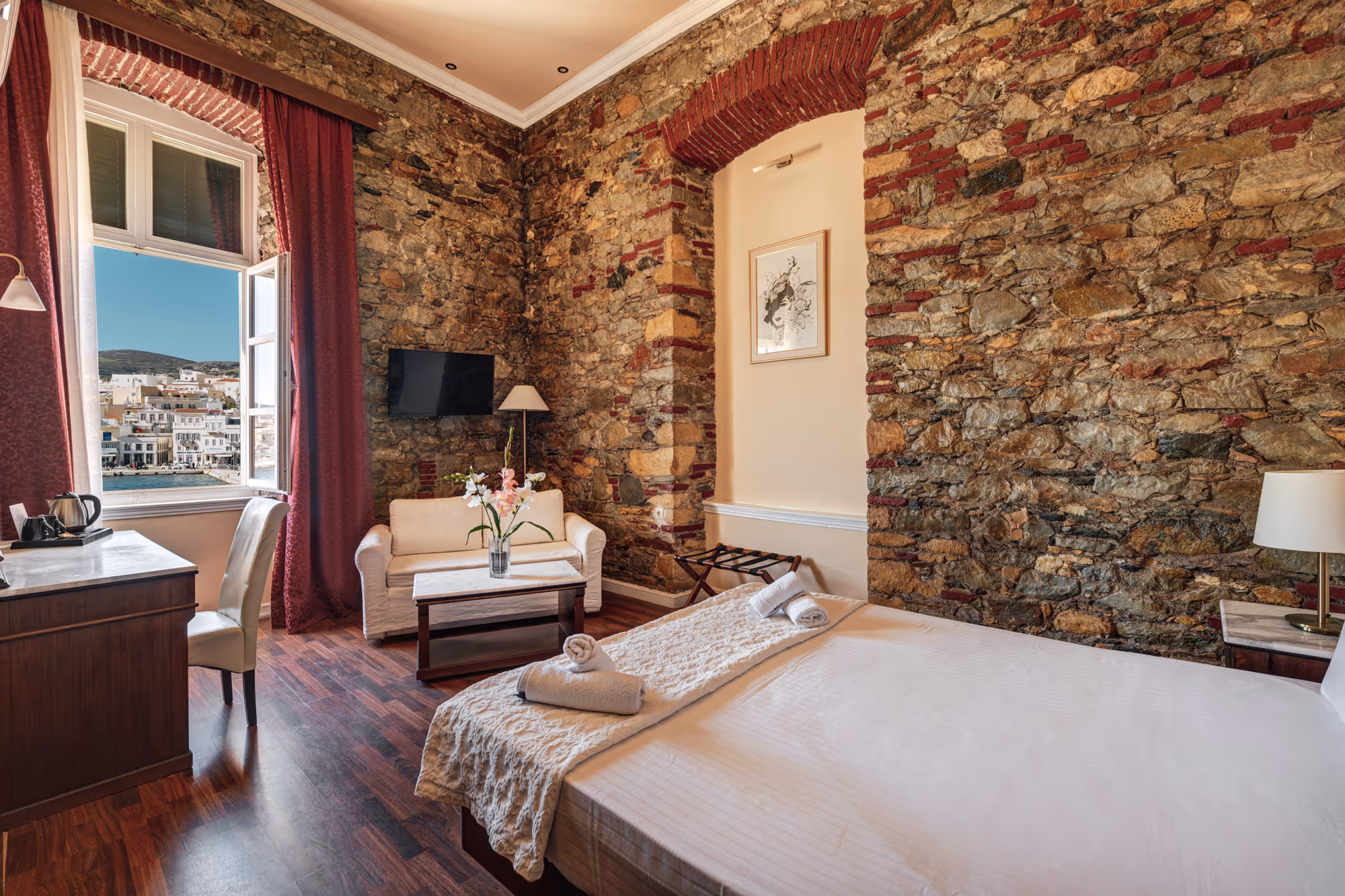 Hotel room with stone walls, a large bed with white linens, a small sofa, a desk, and an open window showing a coastal town view.