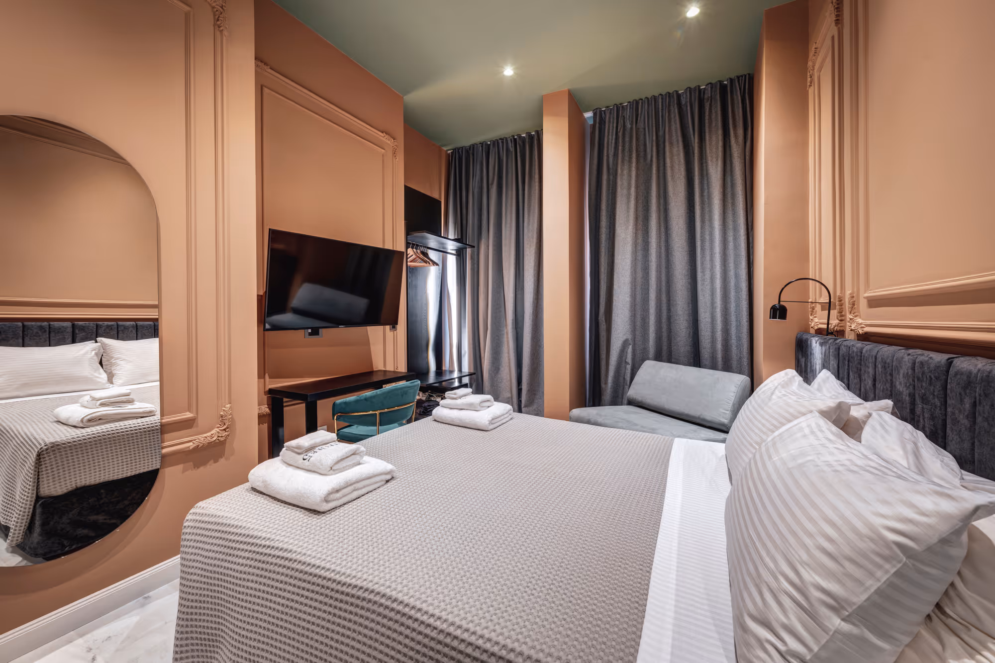 Modern hotel room with a large bed with white pillows and gray blanket, wall-mounted TV, teal chair, gray curtains, and a small gray sofa.