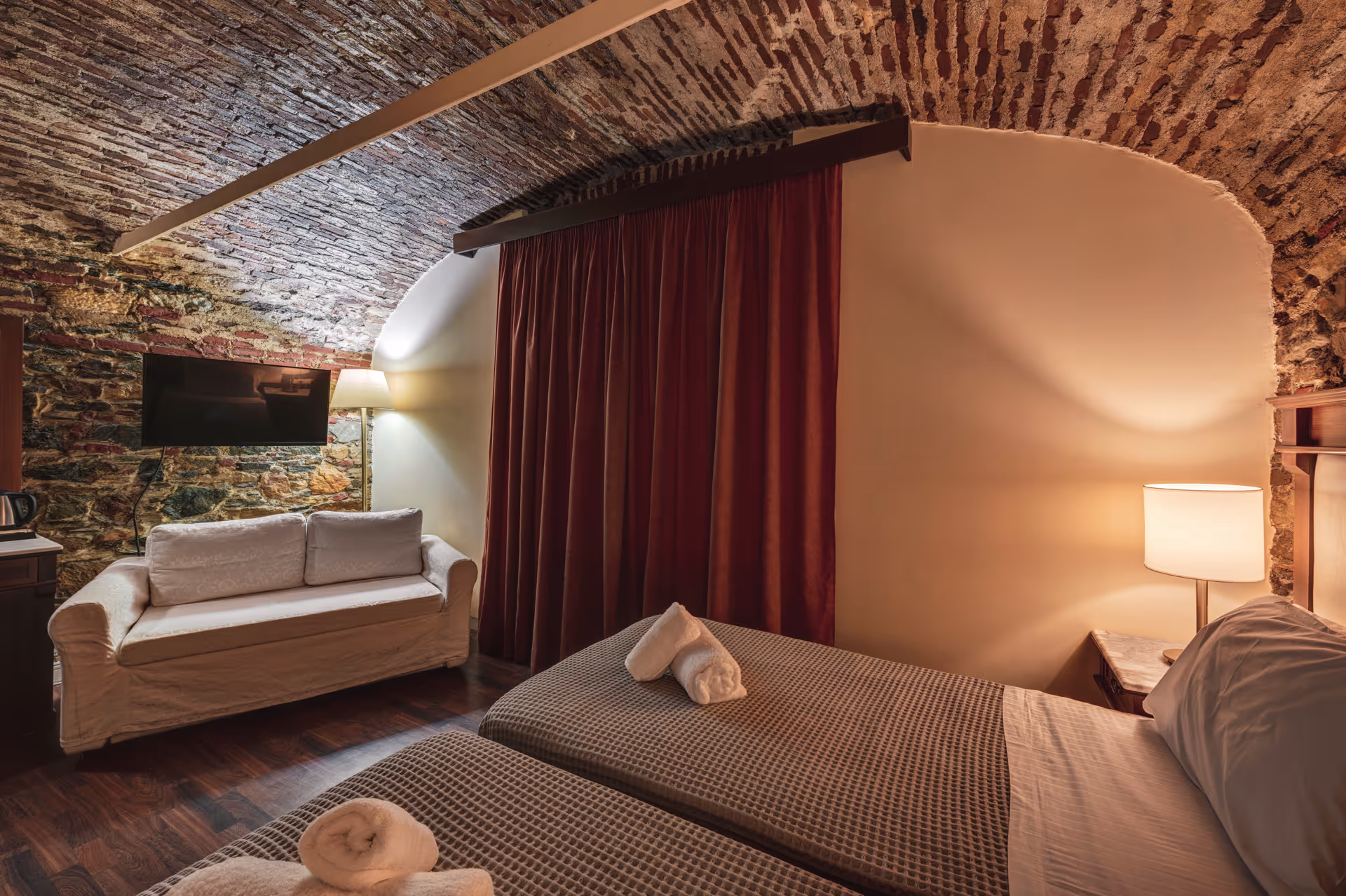 Hotel room with twin beds, white rolled towels, a beige sofa, a wall-mounted TV, and exposed brick vaulted ceiling.