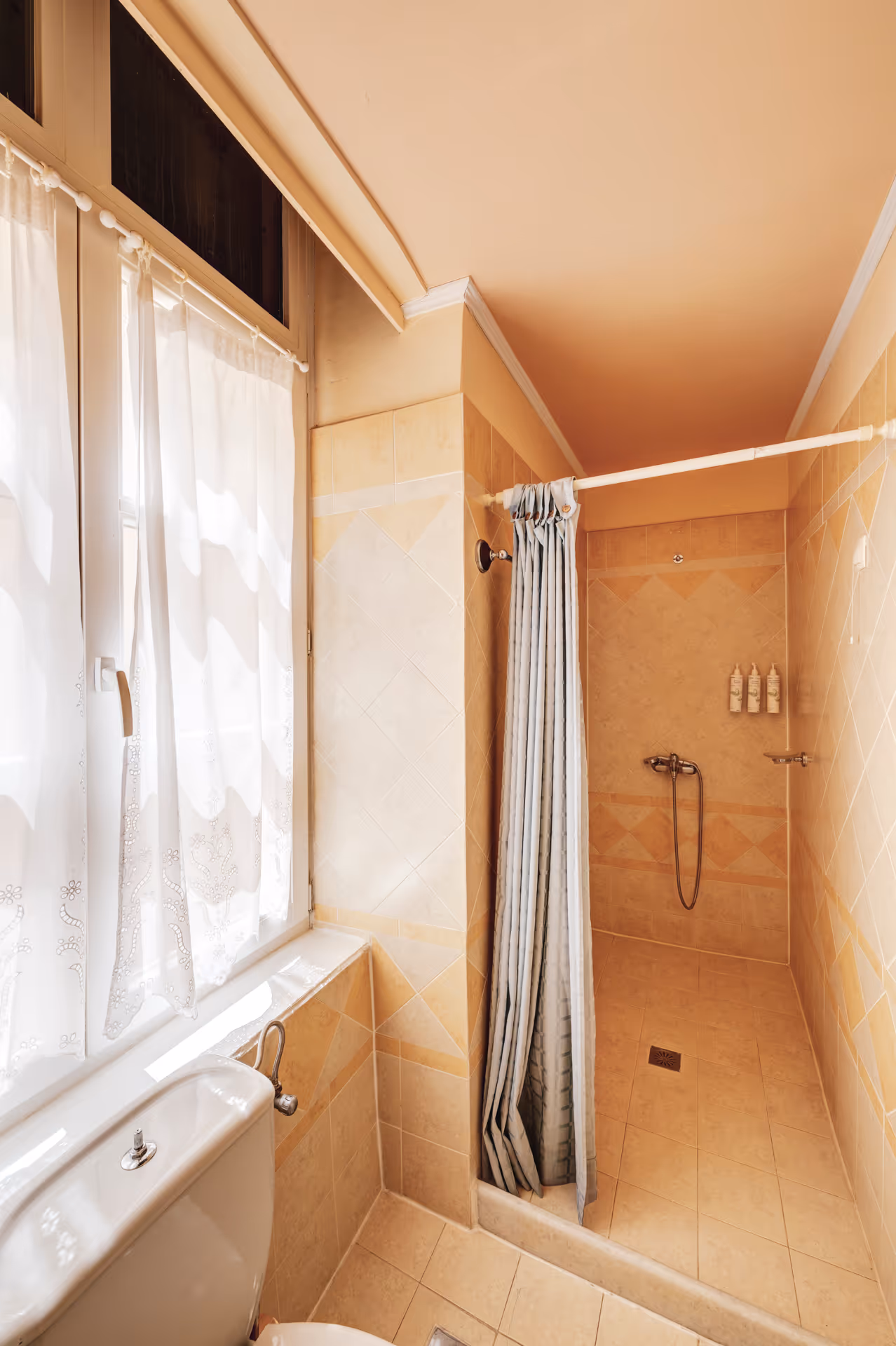 Small bathroom with beige tiled walls and floor, white toilet, sheer window curtains, and a shower area with a closed striped curtain.