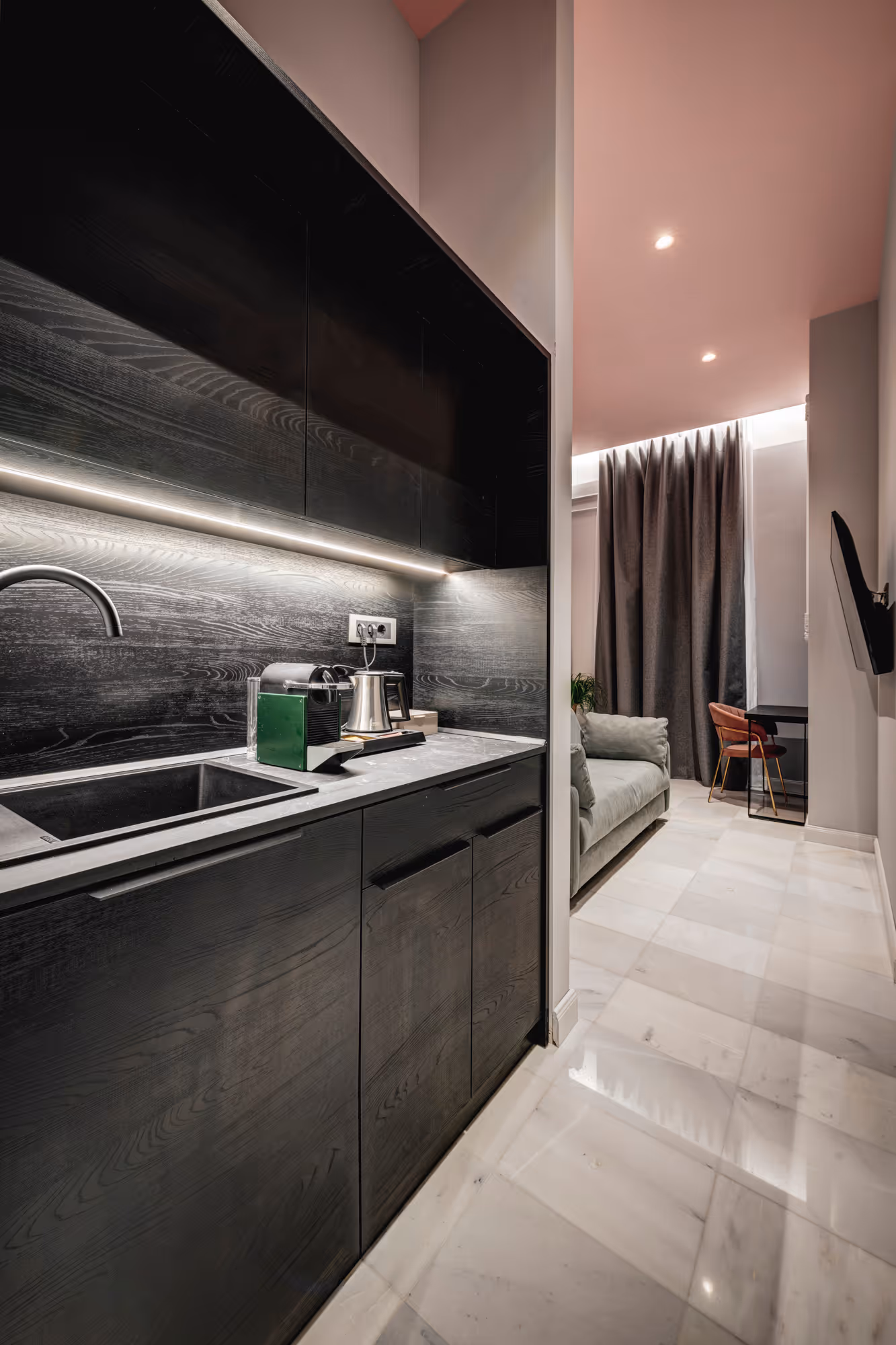 Modern kitchen with black wood cabinets, a sink, coffee machine, and view into a living area with gray sofa and red chair.