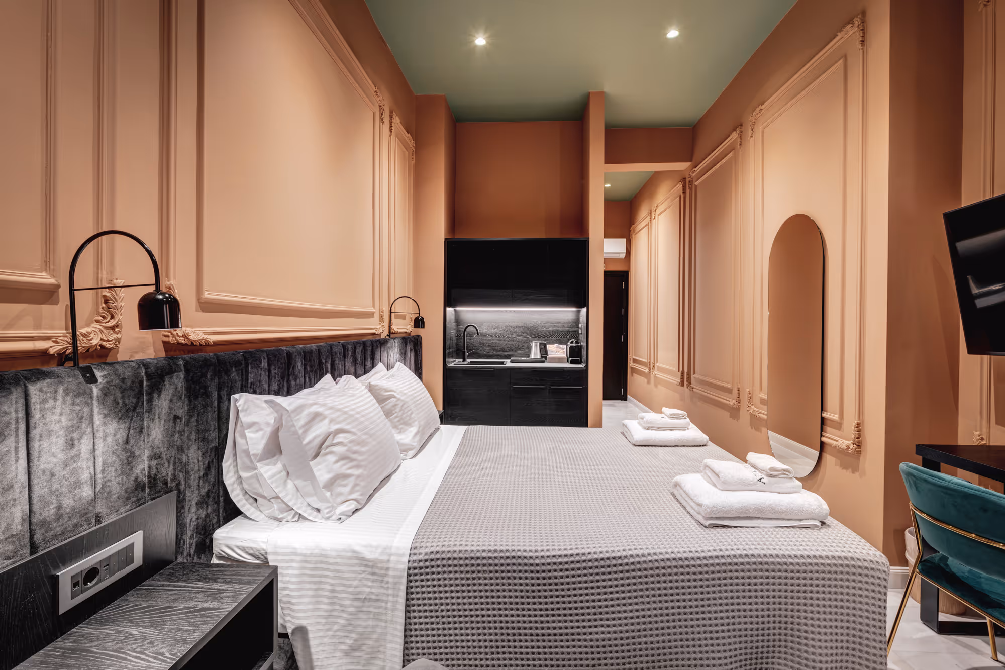Modern hotel room with a large bed featuring white pillows and a gray textured blanket, peach walls, black kitchenette, round mirror, and mounted TV.