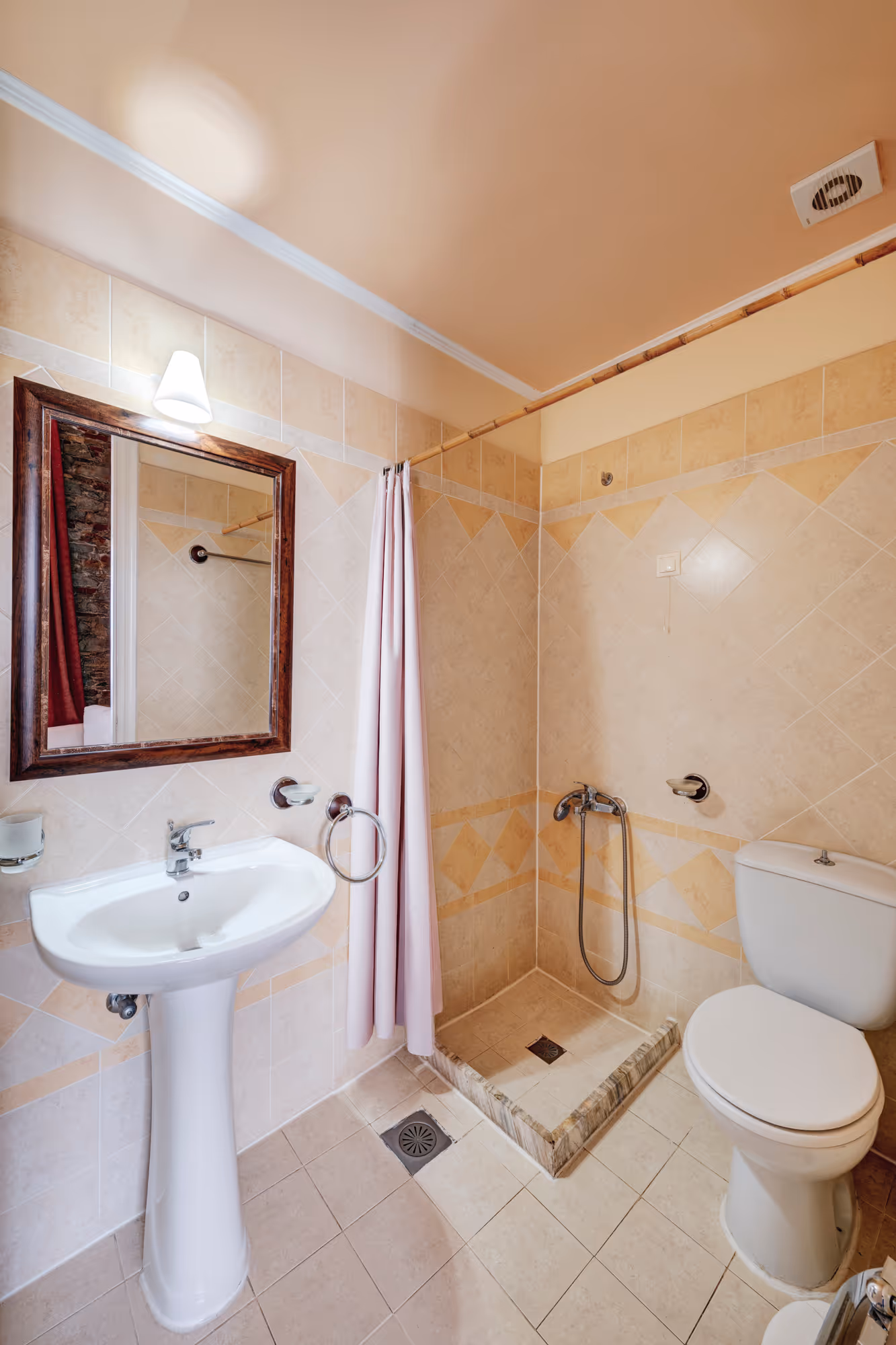 Compact bathroom with beige tiled walls, a pedestal sink under a wooden-framed mirror, a small shower area with a curtain, and a white toilet.